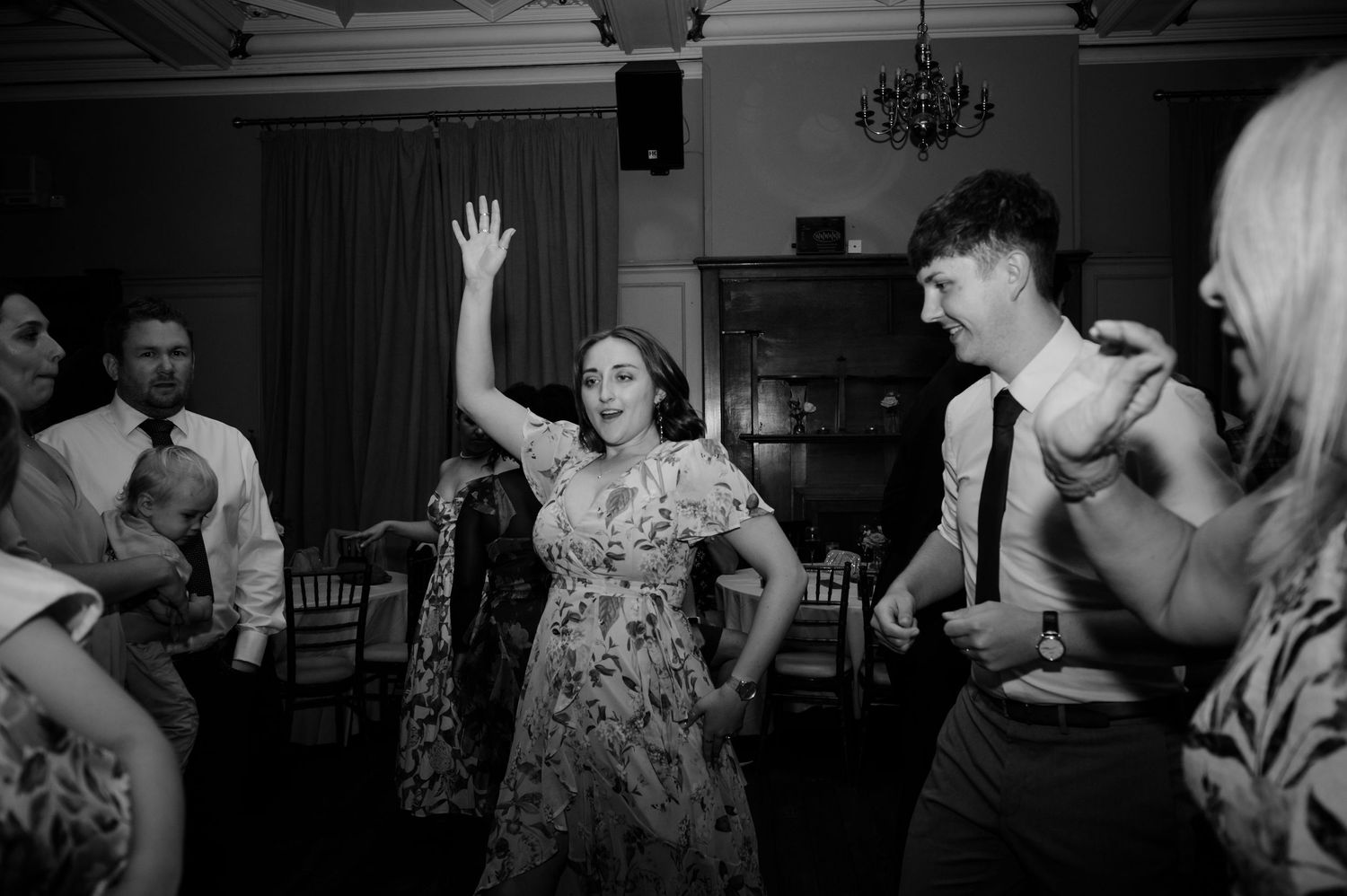 Black and white photo of a woman raising her arm and dancing joyfully with guests on a crowded wedding dance floor.
