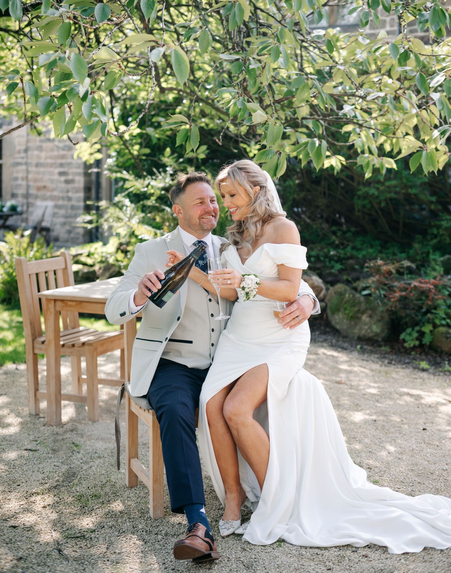 Bride in an off-shoulder white gown and groom in a gray suit toast with champagne on a wooden bench in a sunny garden.
