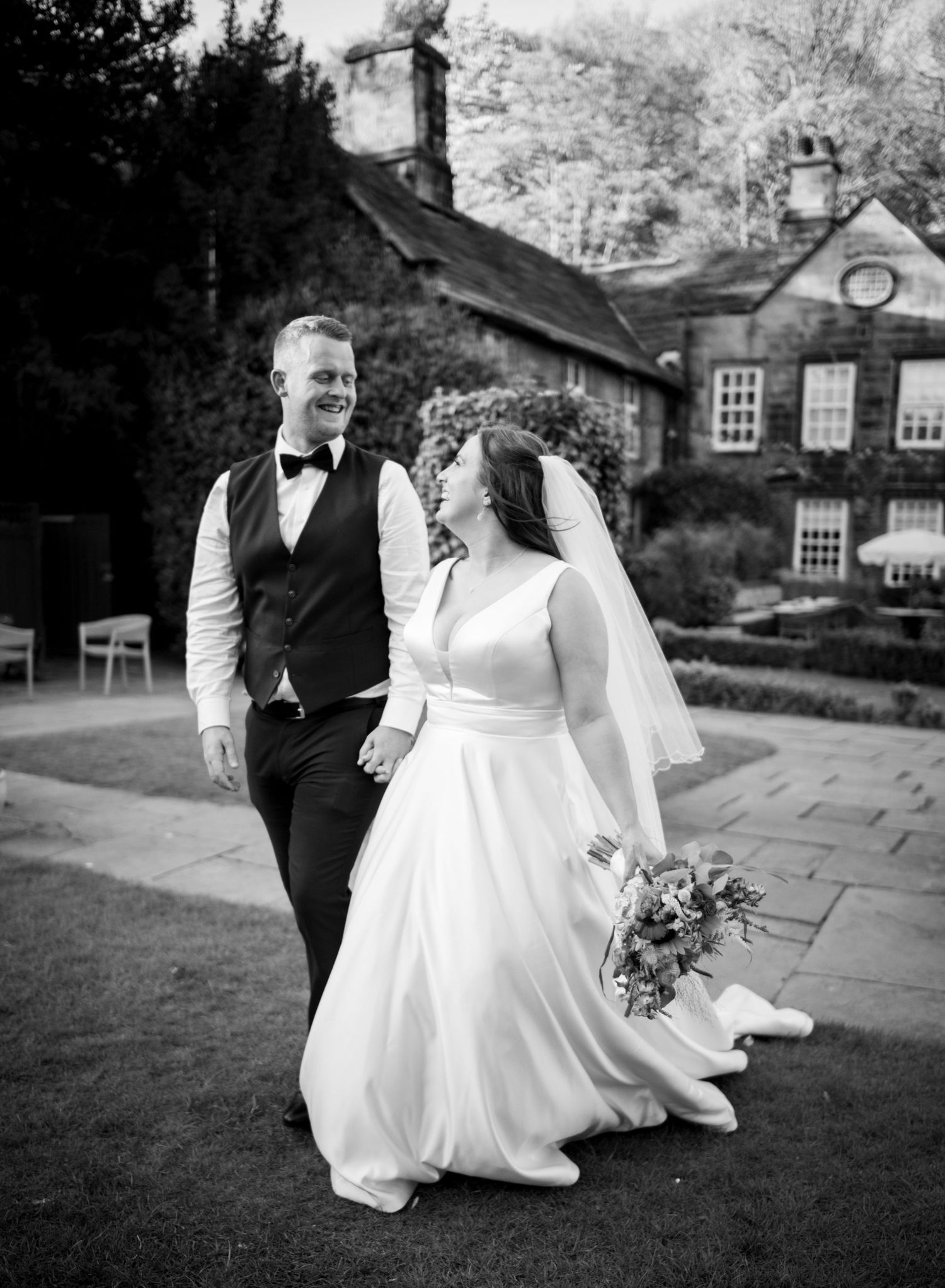Black and white photo of a smiling bride and groom walking hand in hand outside a historic stone manor house.
