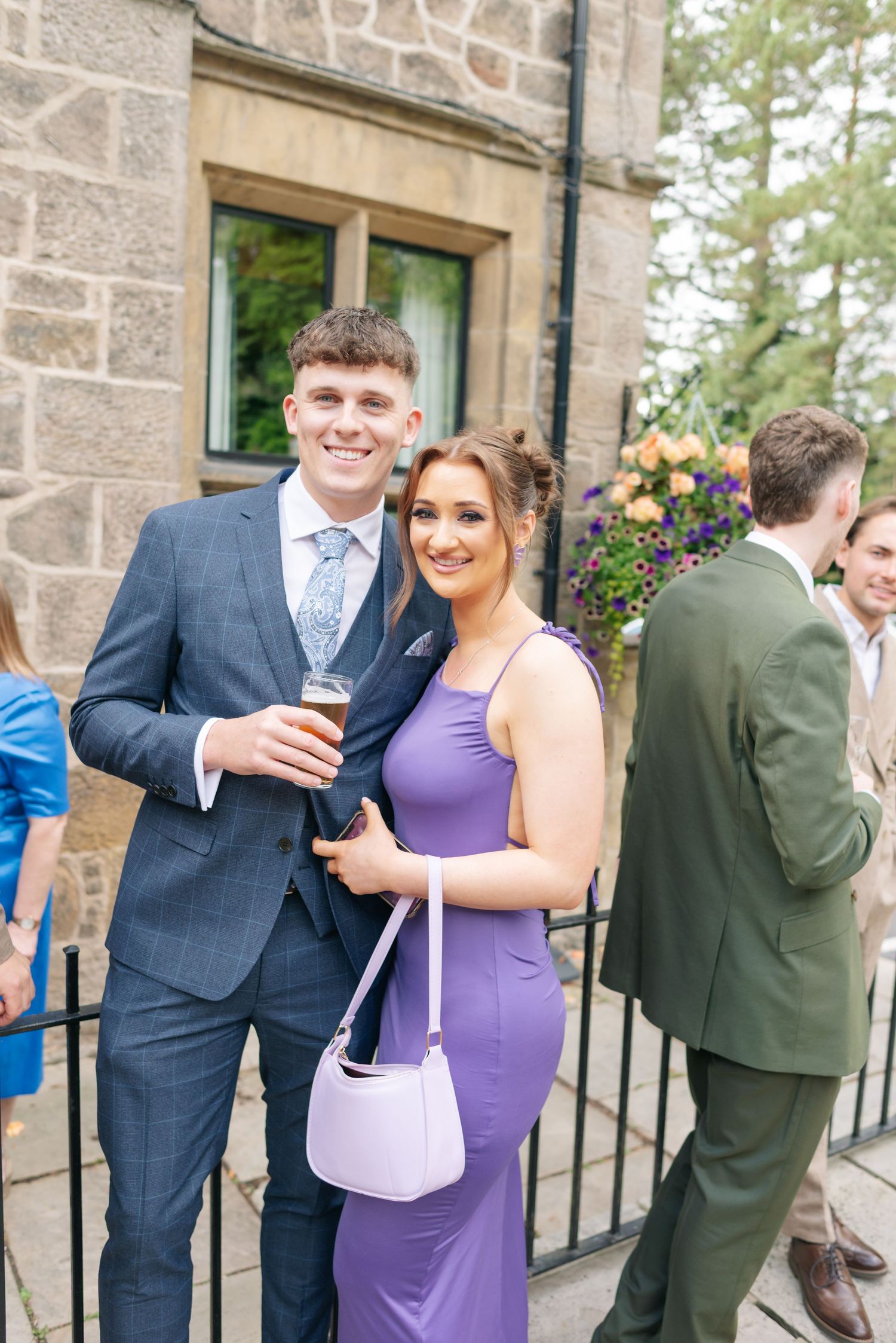 Wedding guests in formal attire mingle and socialize outside a stone building during a reception.