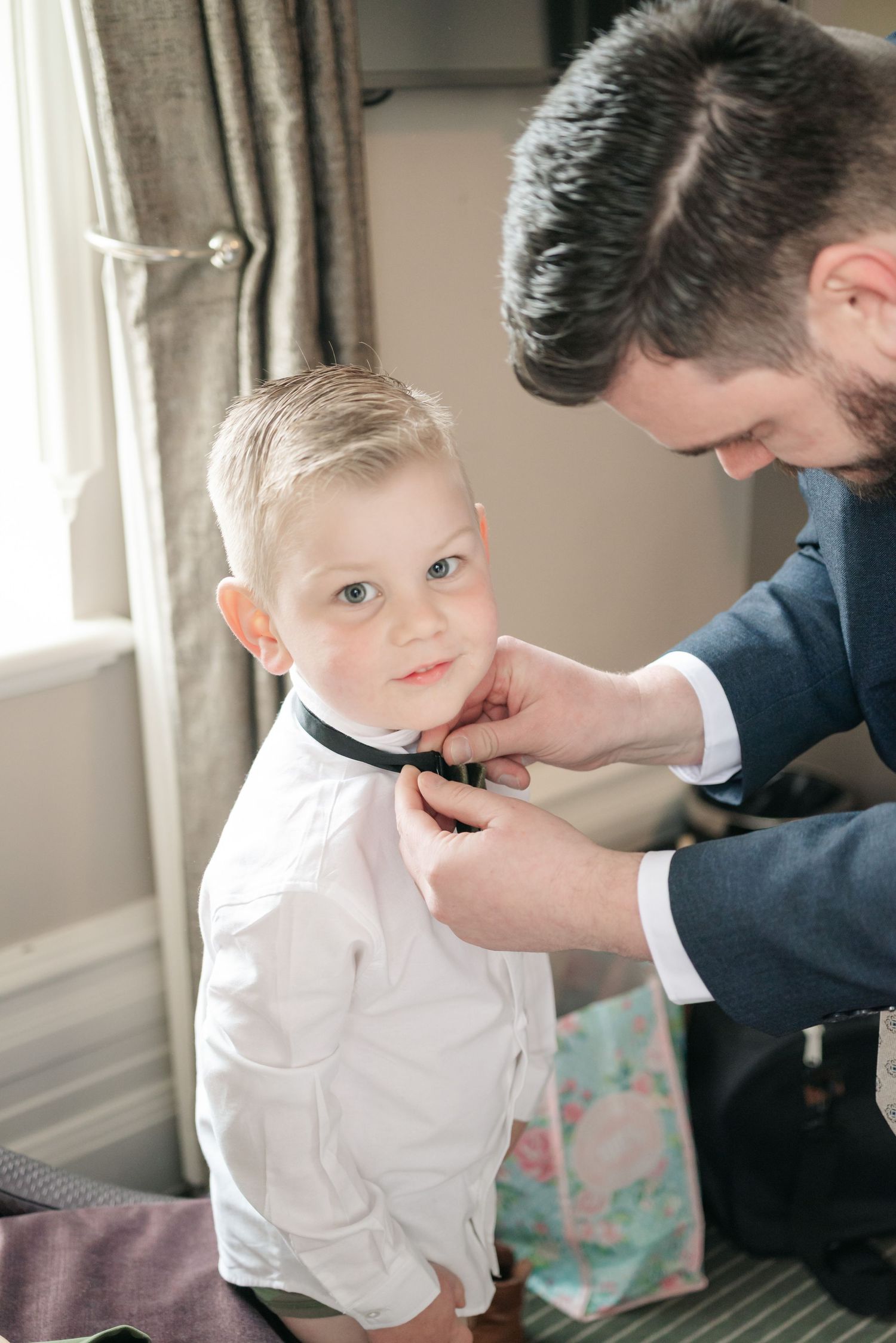 A young boy in a white dress shirt gets help with his bow tie during formal preparation.