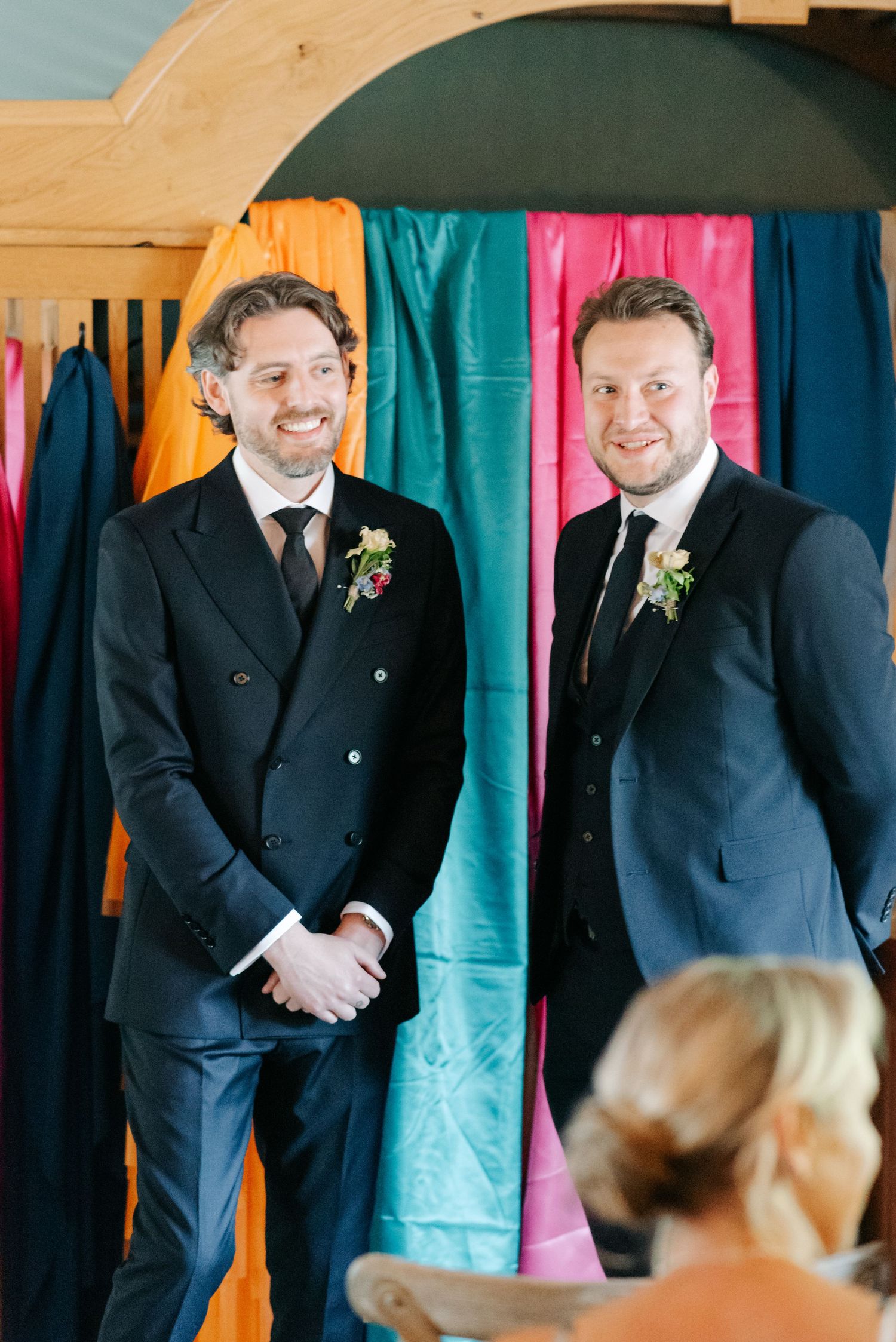 Groomsmen in black suits standing against a backdrop of colorful fabric panels.