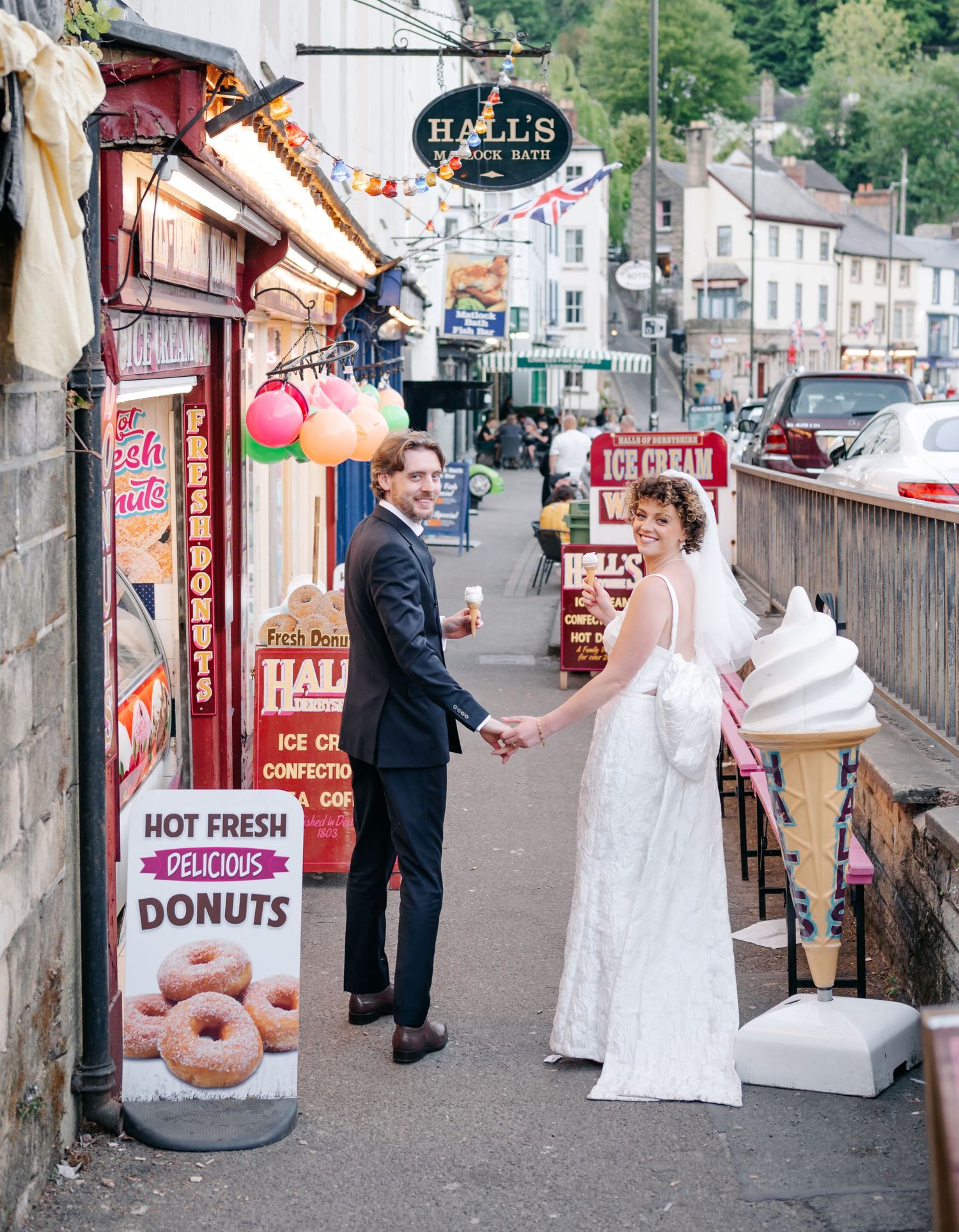 A couple in wedding attire poses for photos in front of a donut shop storefront.
