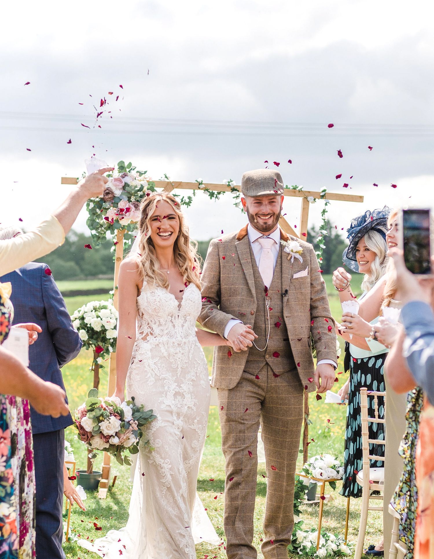 Wedding couple walks down aisle as guests shower them with red rose petals during outdoor countryside ceremony.
