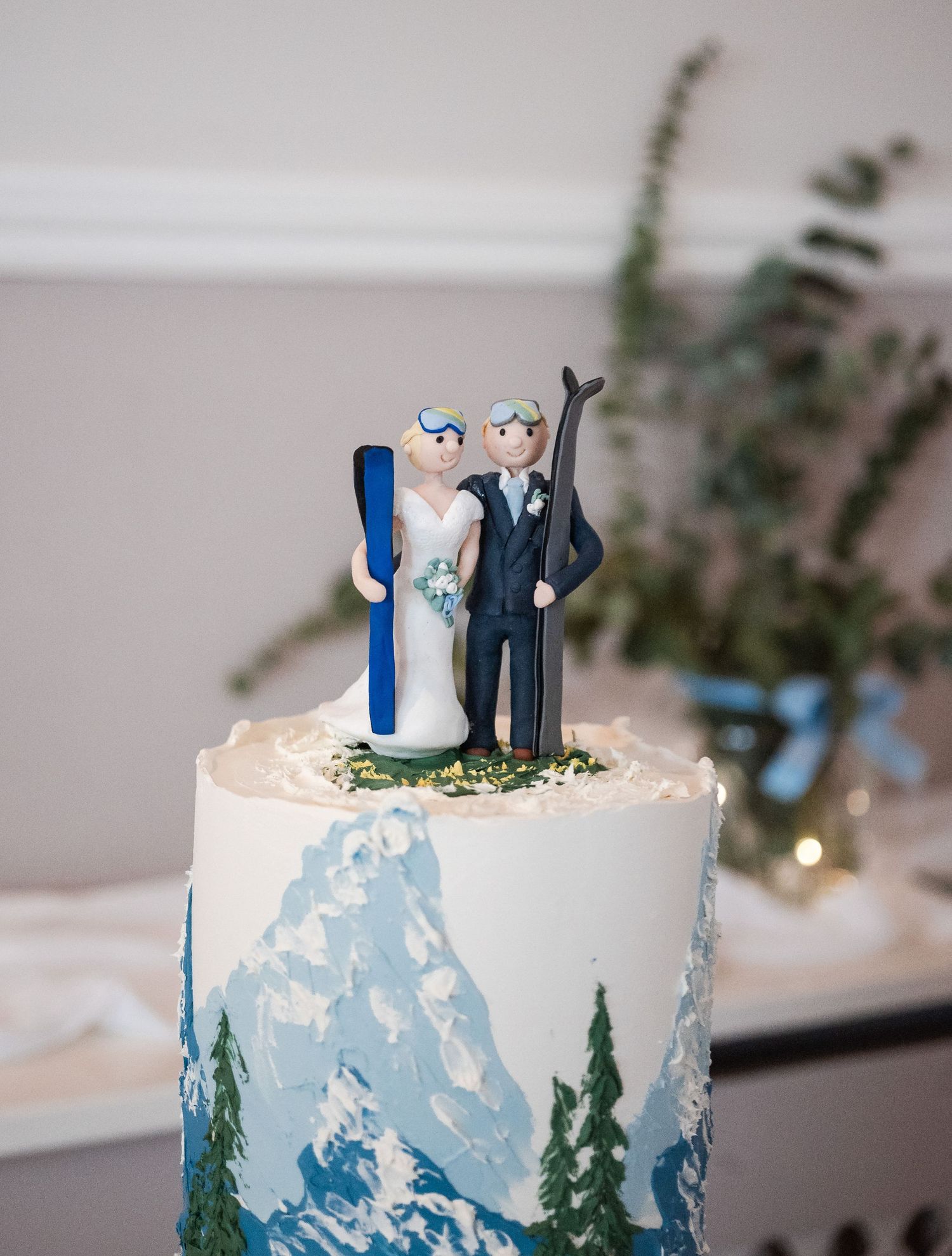 White wedding cake decorated with blue mountain scenery and miniature couple topper.