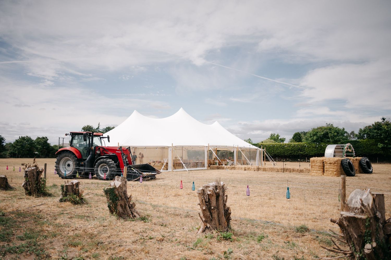 A white wedding tent set up in a rural field with red and green tractors parked nearby under a cloudy sky.