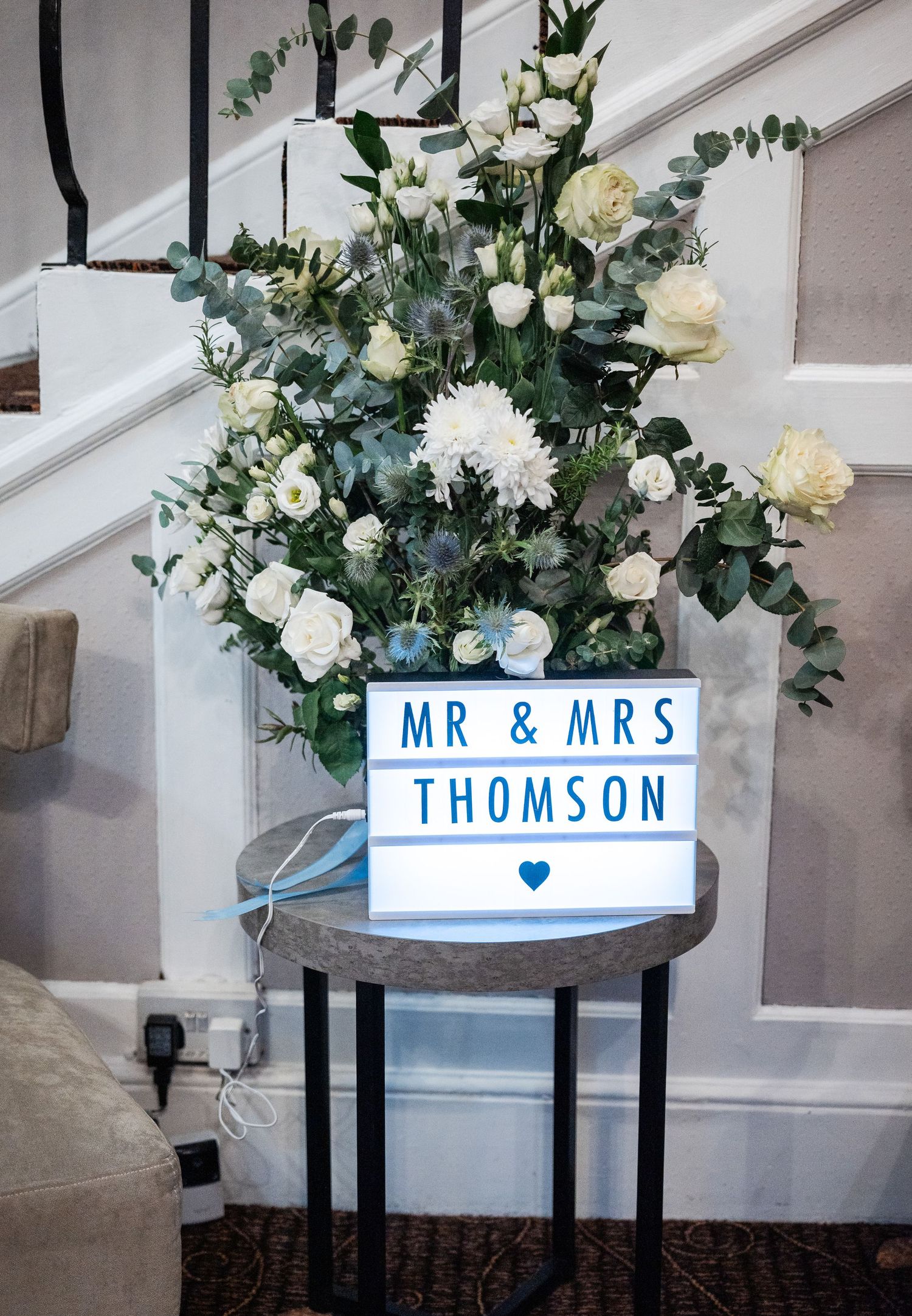 Elegant white floral arrangement with a Mr & Mrs Thomson lightbox sign against grey stairs.