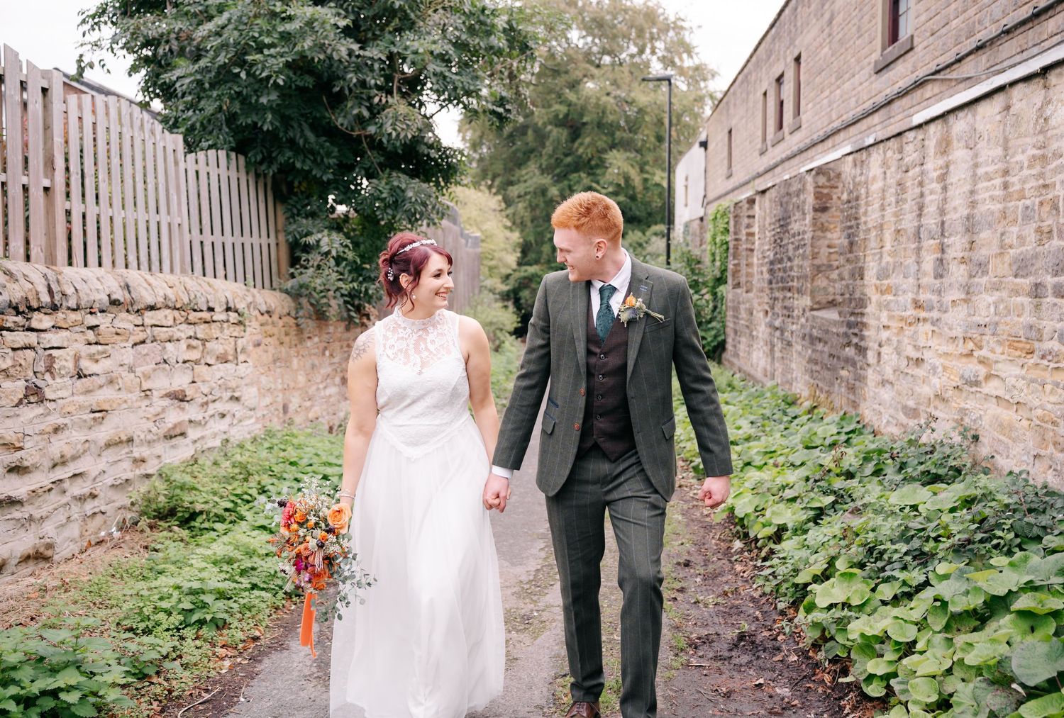 Couple walking hand in hand down a stone-walled pathway in wedding attire.