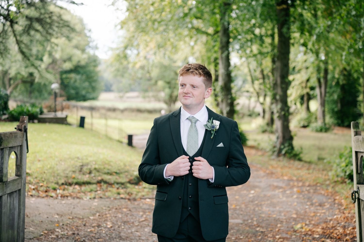 A wedding party walks down a tree-lined autumn path with fallen leaves on the ground.