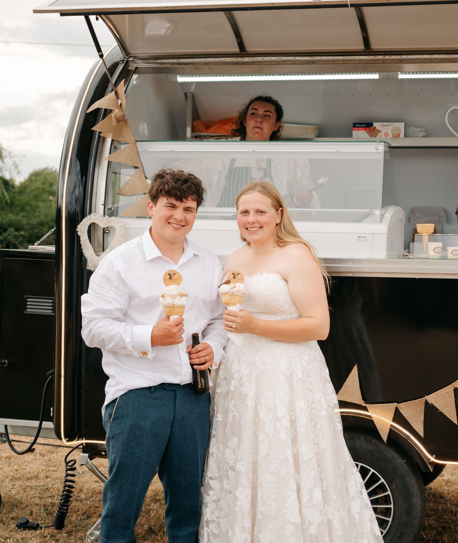 Newlyweds enjoy ice cream treats together near vintage food truck at outdoor reception.