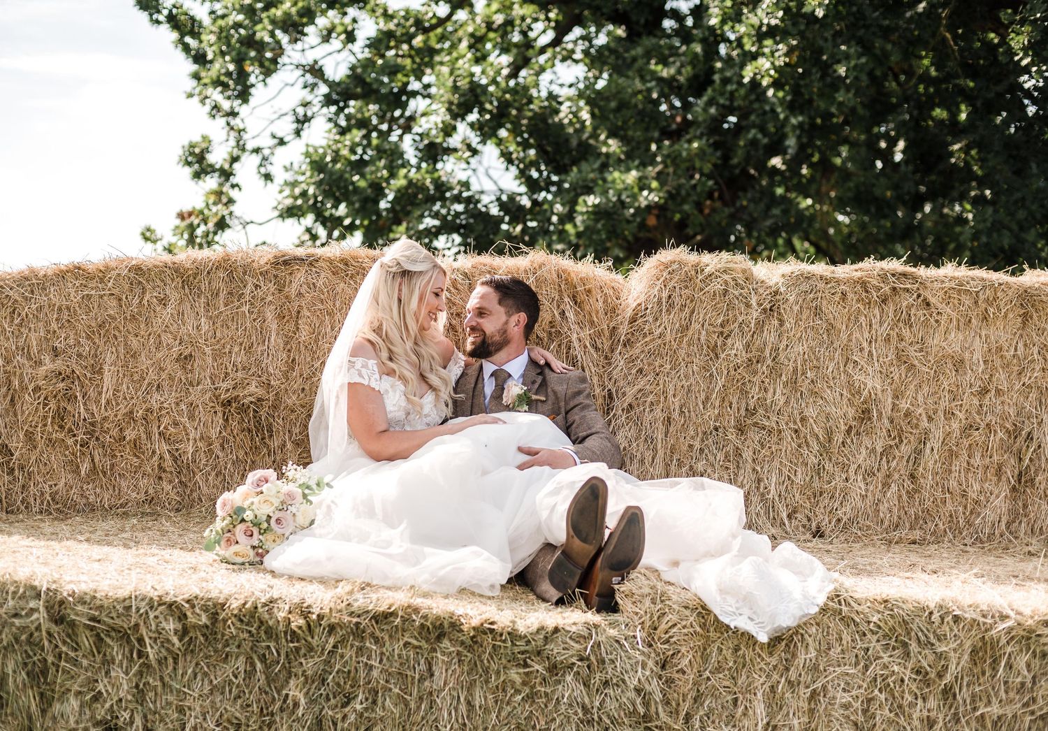 Newlyweds relaxing on hay bales during rustic outdoor photoshoot.