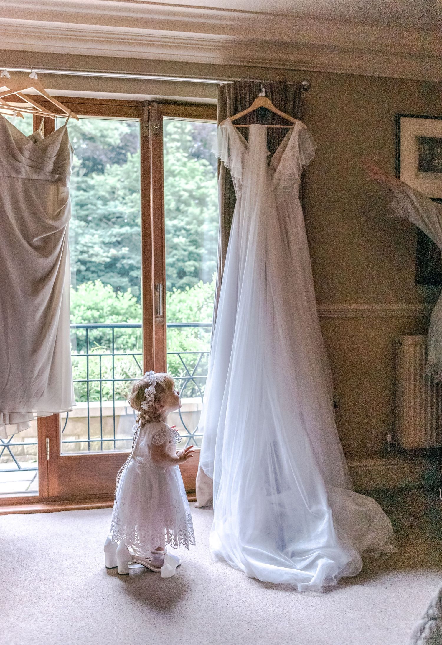 A wedding dress hangs by a window while a small flower girl in white lace looks up at it.