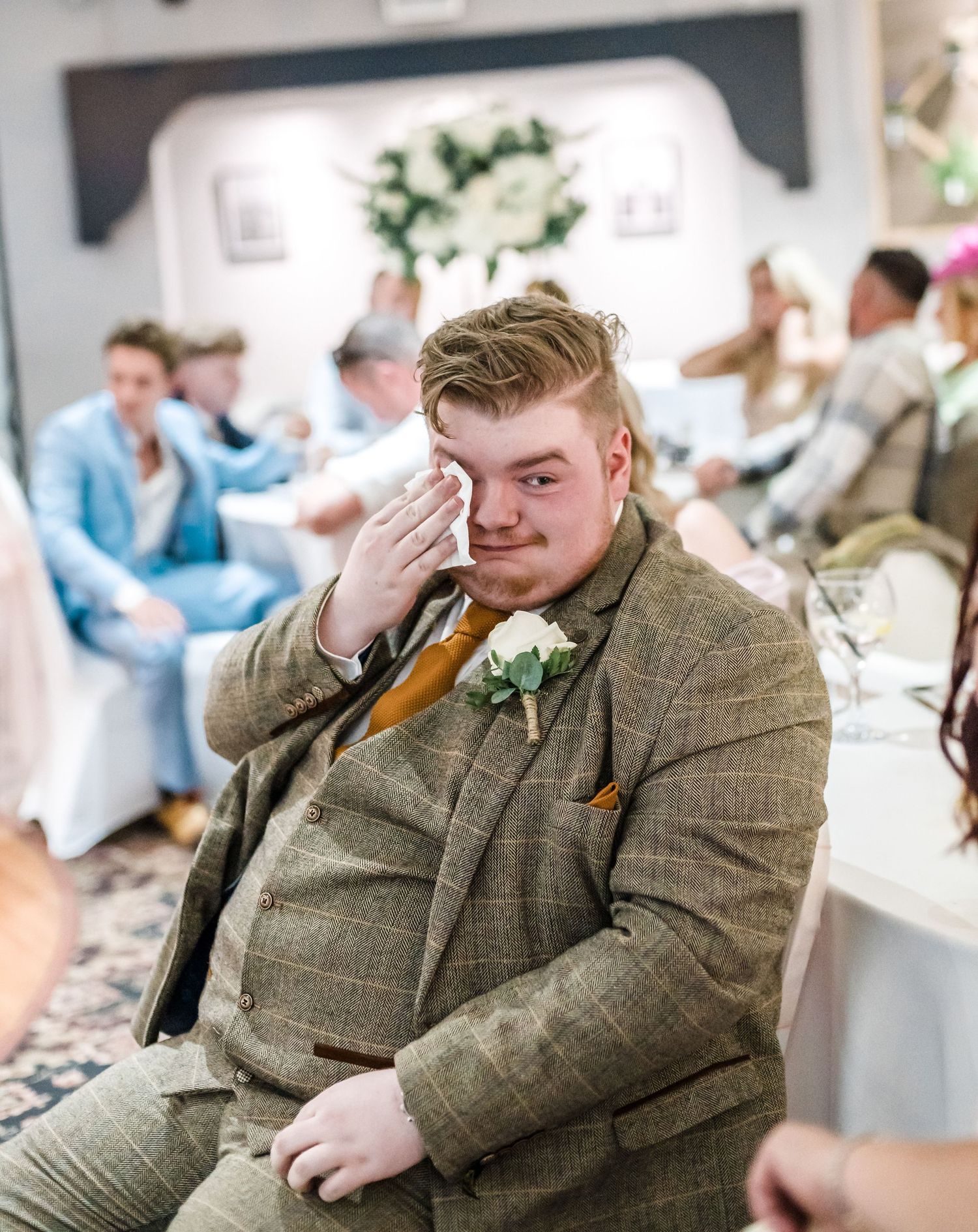Wedding guest in tweed suit reacts emotionally during reception speeches.