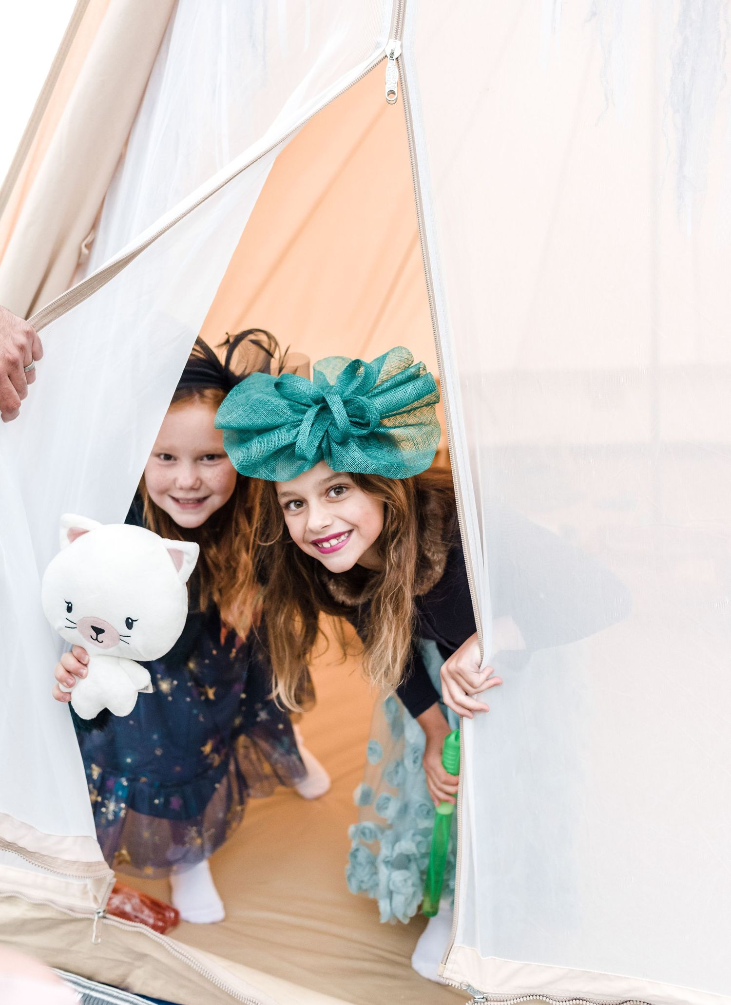 Two children peek playfully from a white tent with one wearing a teal bow.