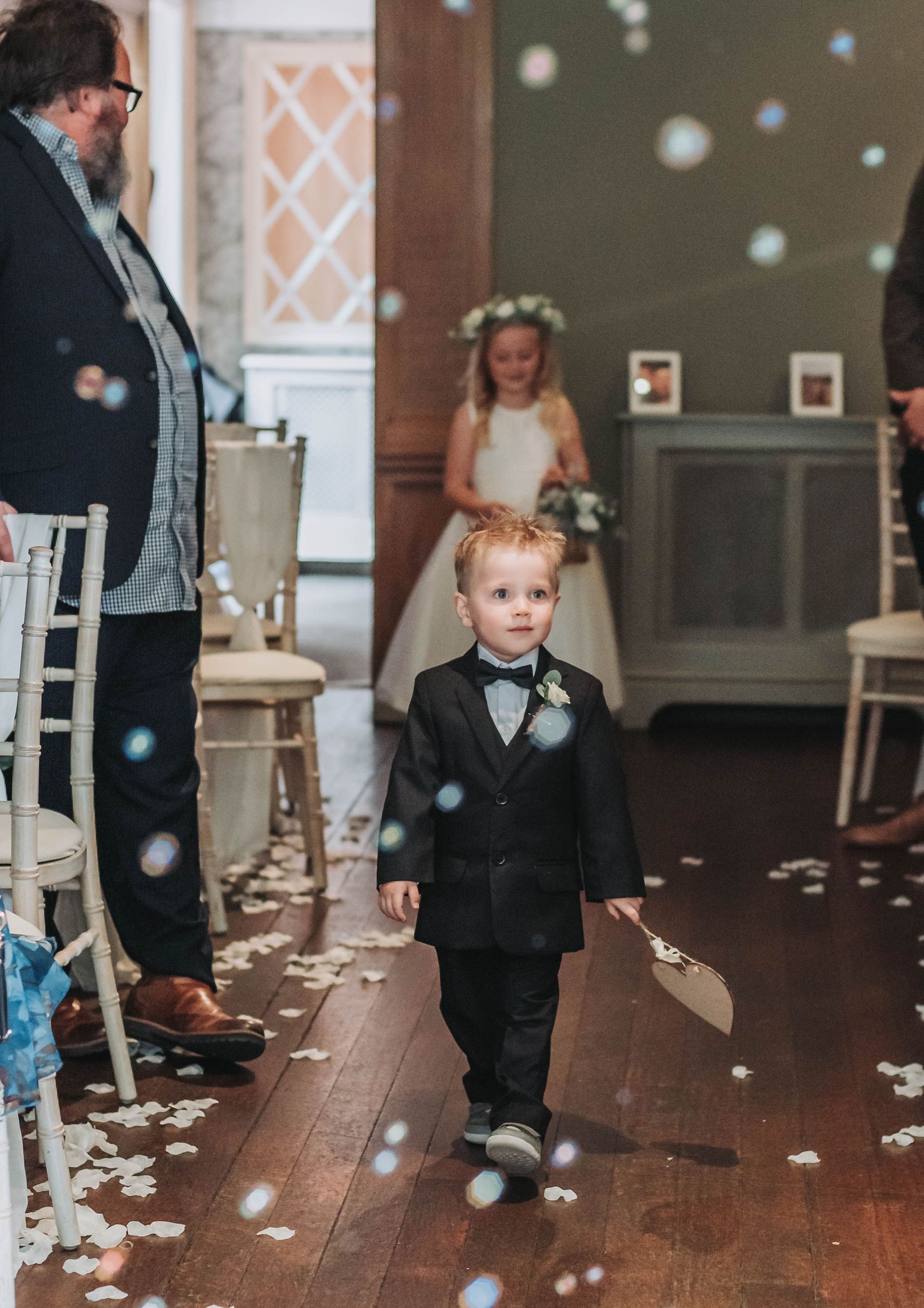 A young ring bearer in black tuxedo walks down the aisle with flower petals scattered around.