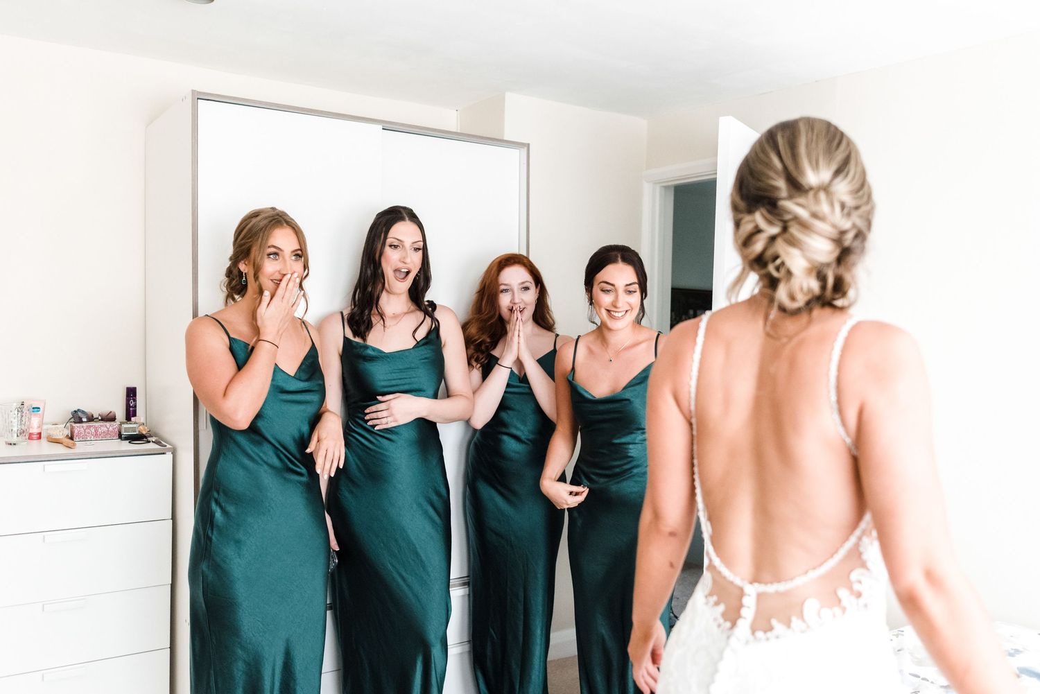 Bridesmaids in emerald dresses react with joy to seeing the bride in her backless wedding gown.