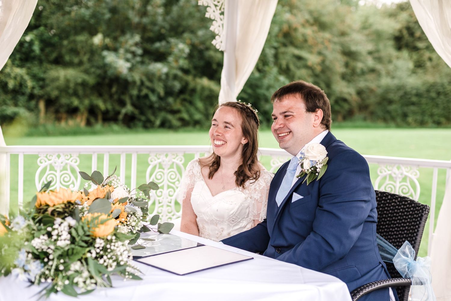 Couple shares a laugh while signing marriage documents under white ceremony arch.