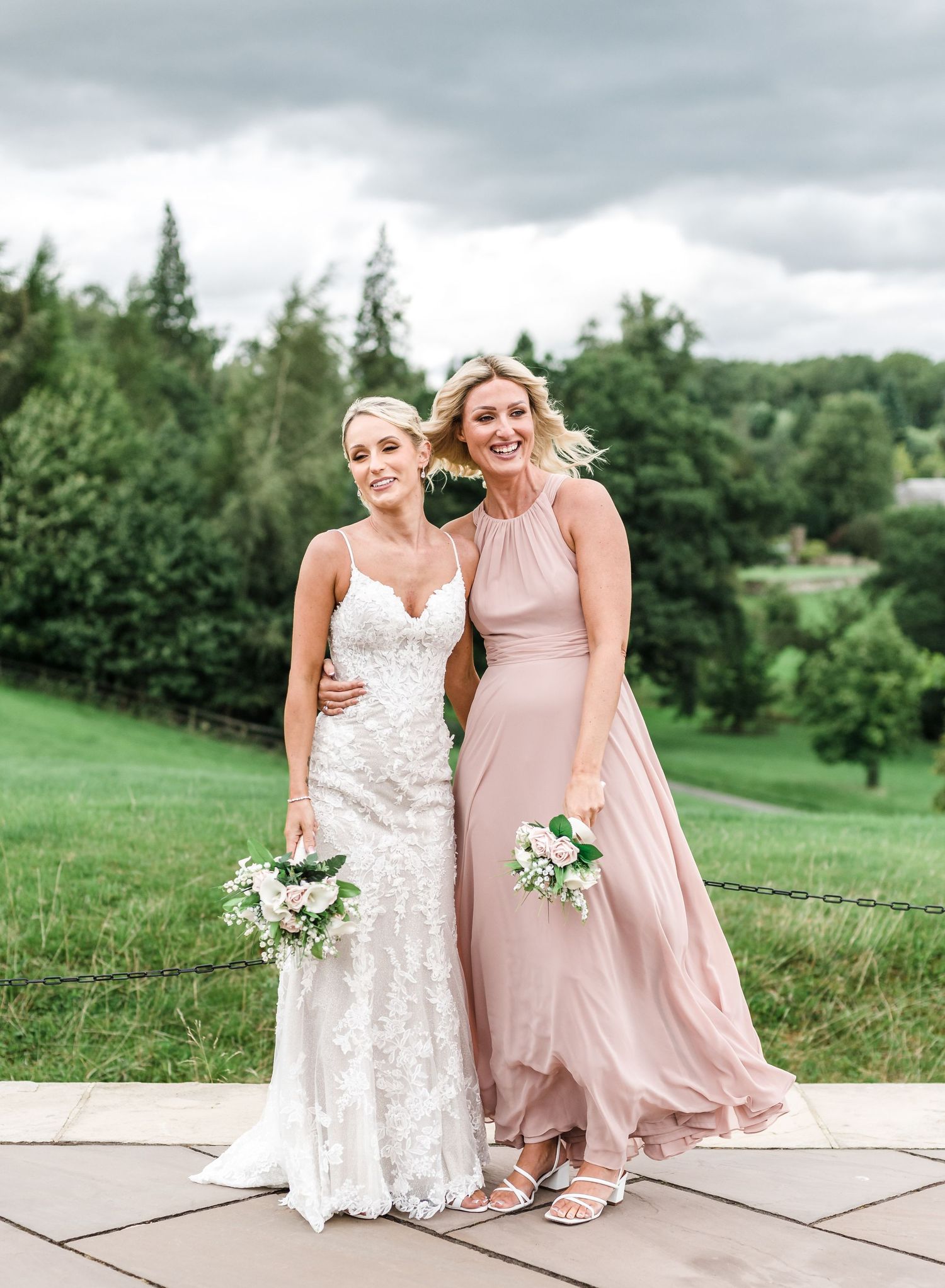 Bride in lace gown and bridesmaid in blush dress pose together outdoors.