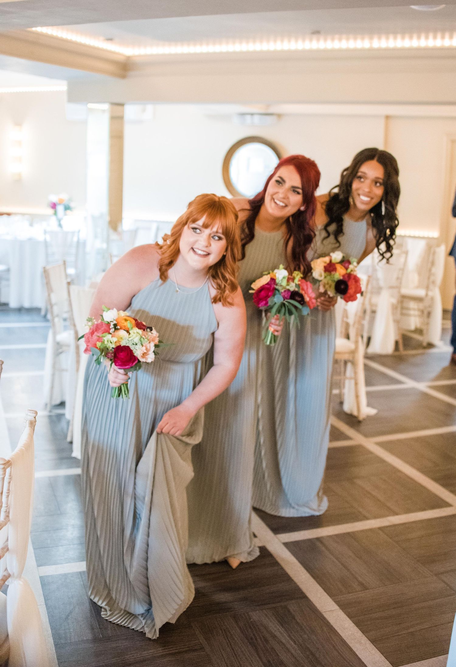 Bridesmaids in flowing grey dresses pose together holding colorful bouquets.