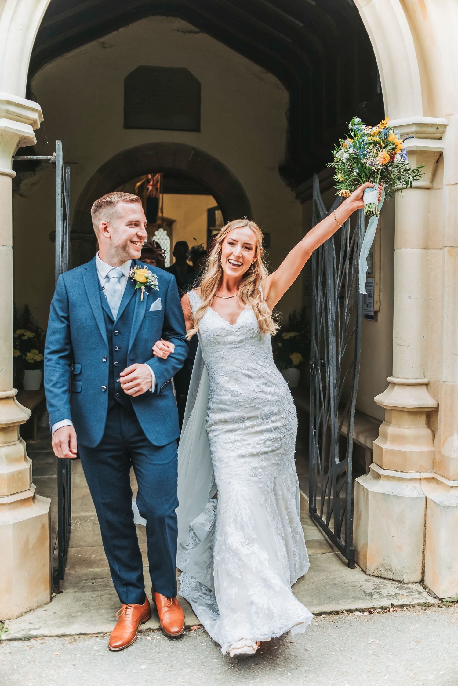 Joyful newlyweds exit a stone archway with the bride raising her bouquet in celebration.