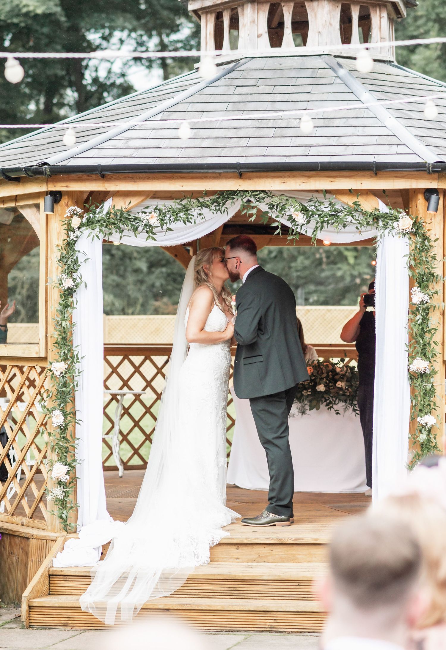 Wedding ceremony in decorated gazebo with greenery garland and white fabric draping.