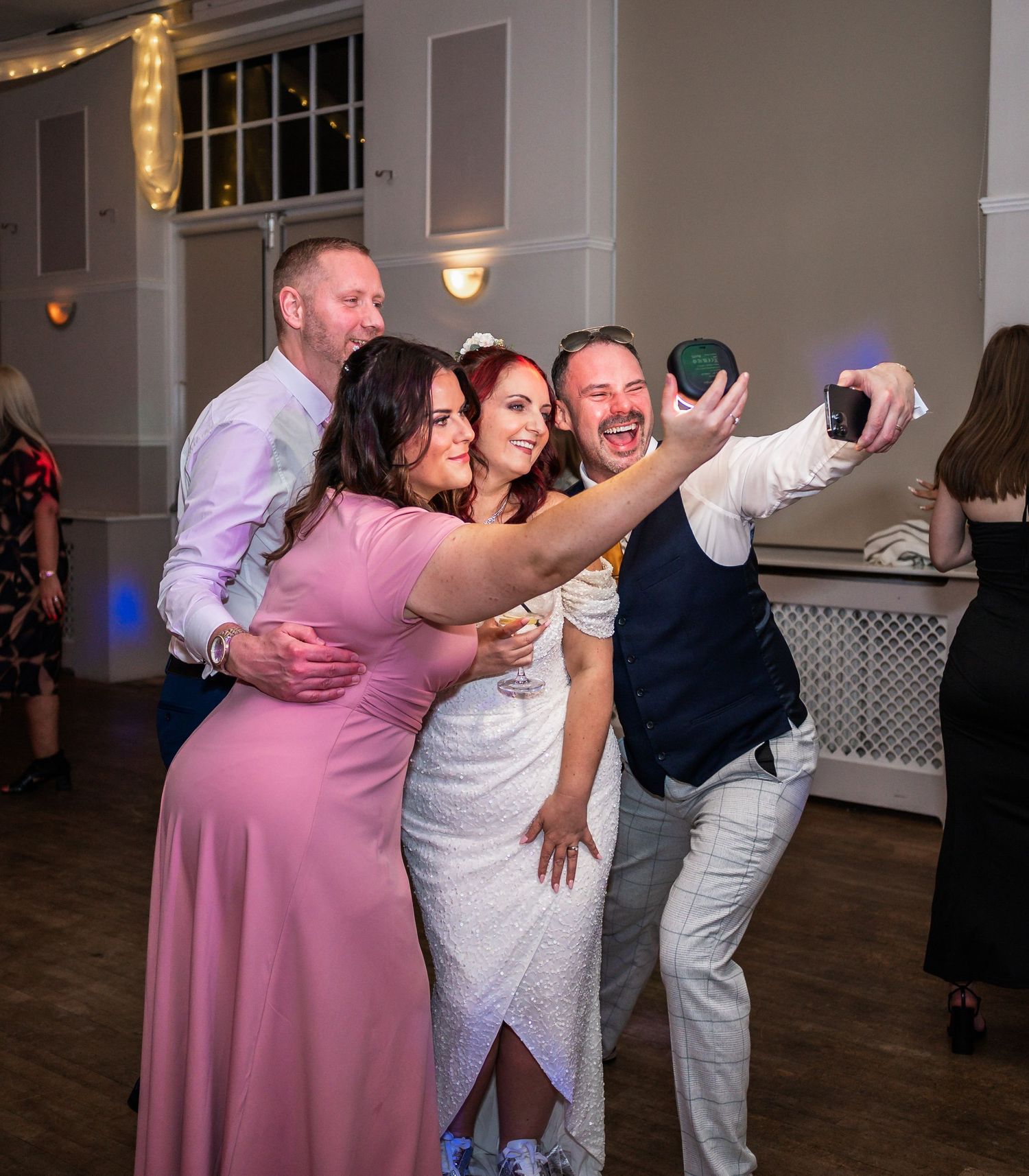 Guests taking celebratory selfies at a wedding reception on a wooden dance floor.