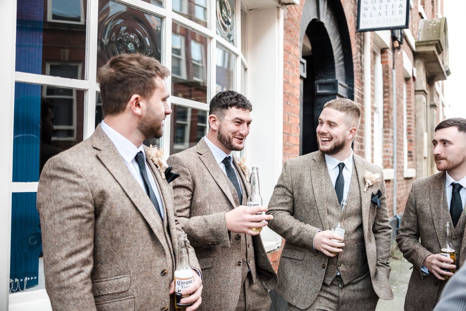 Groomsmen in tweed suits share laughs outside historic building.