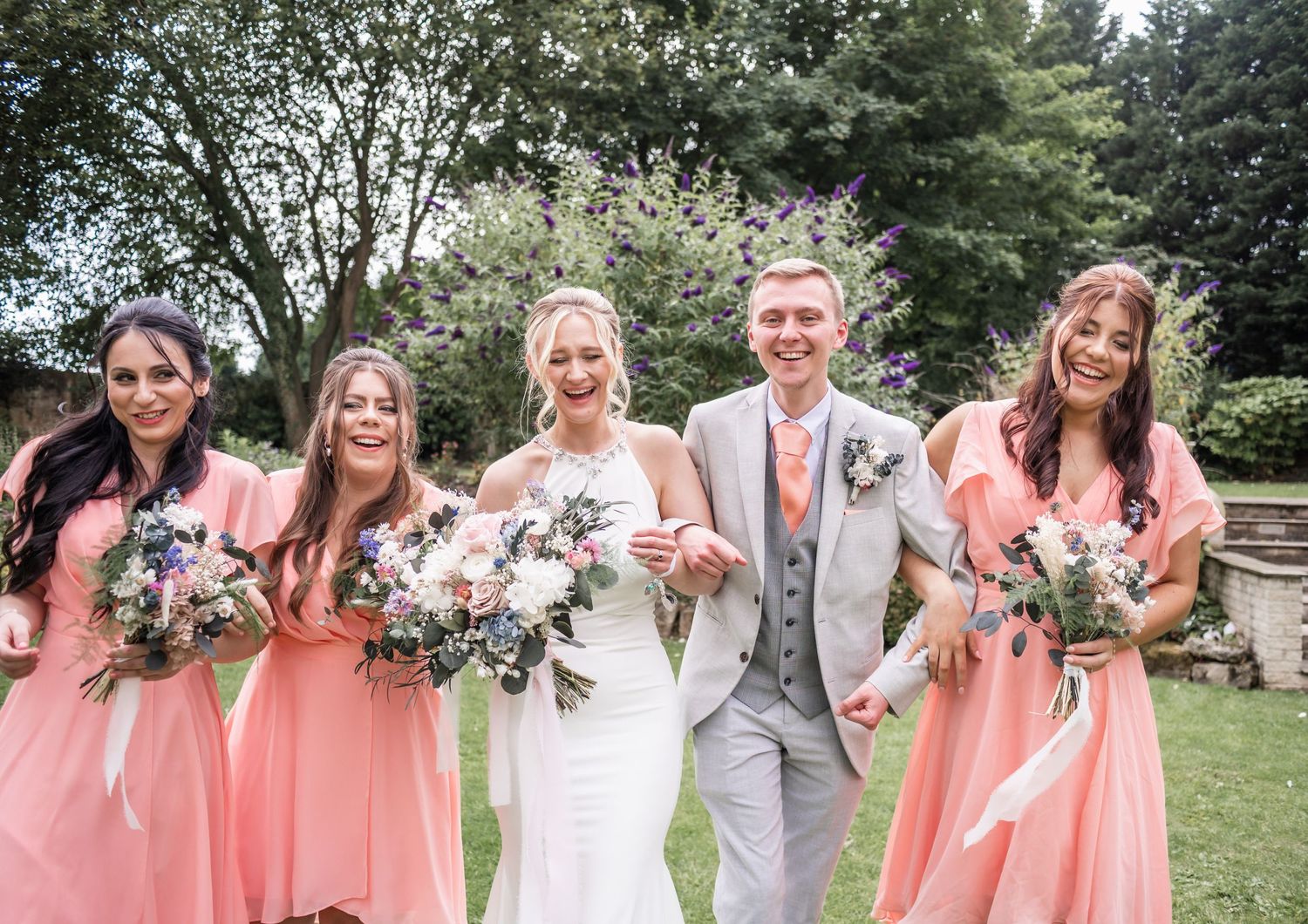 Wedding party in coral dresses and grey suit smile together outdoors.