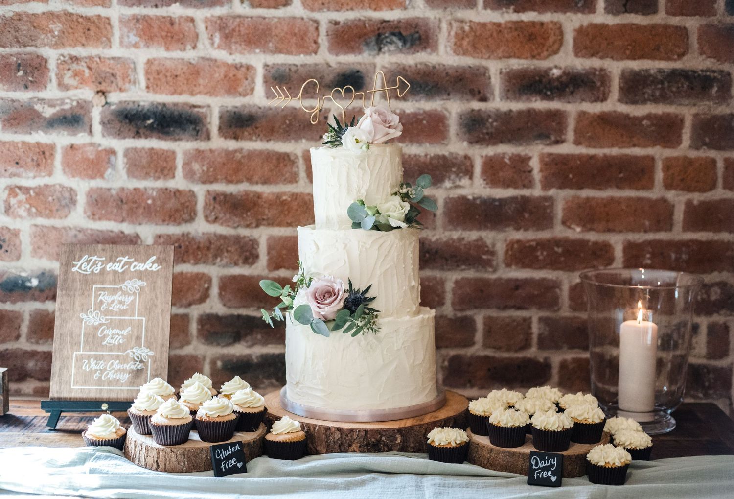 White tiered wedding cake decorated with fresh flowers against rustic brick wall.