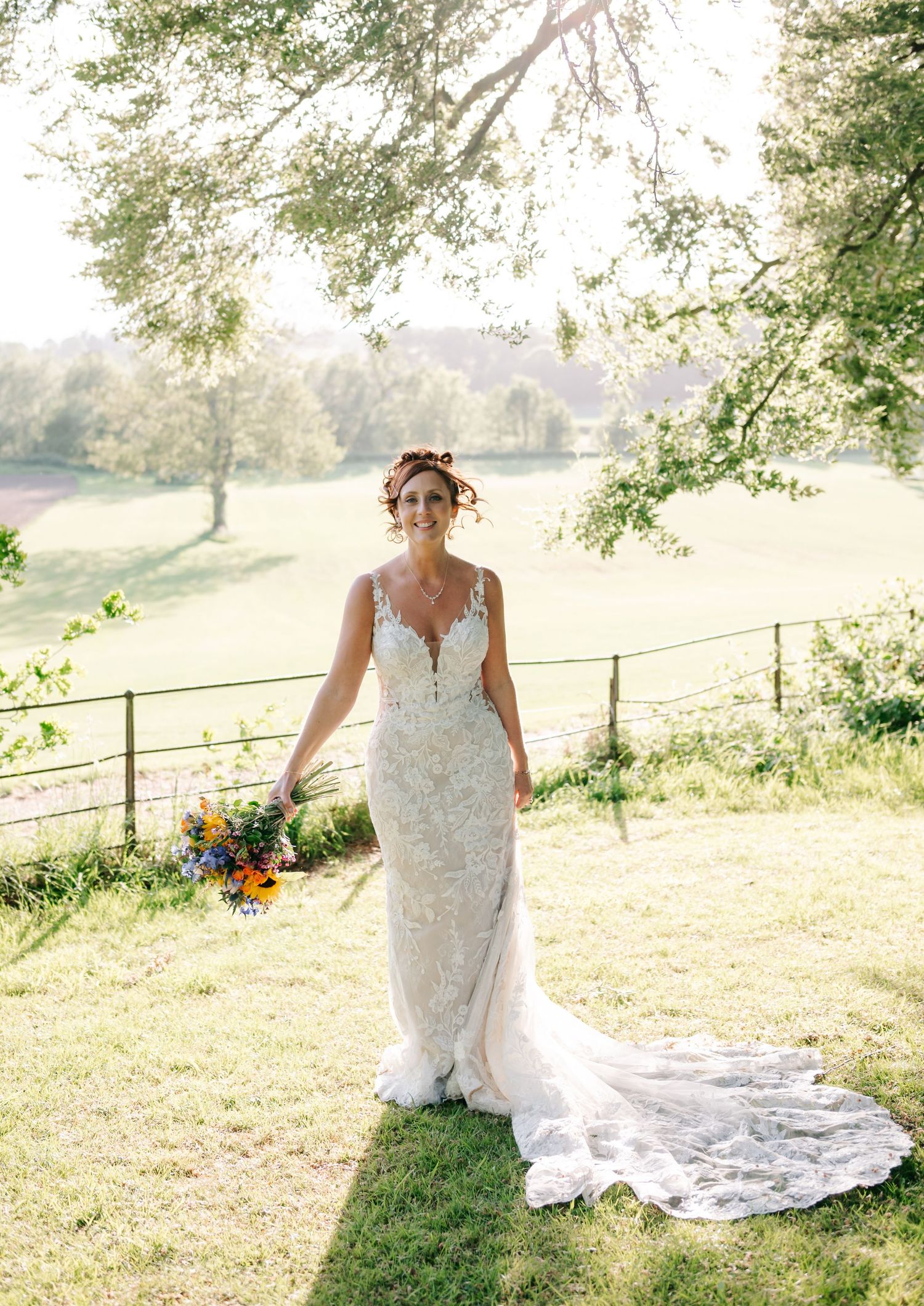 Glowing sunlit portrait of bride in lace gown with long train on grassy lawn under overhanging trees.