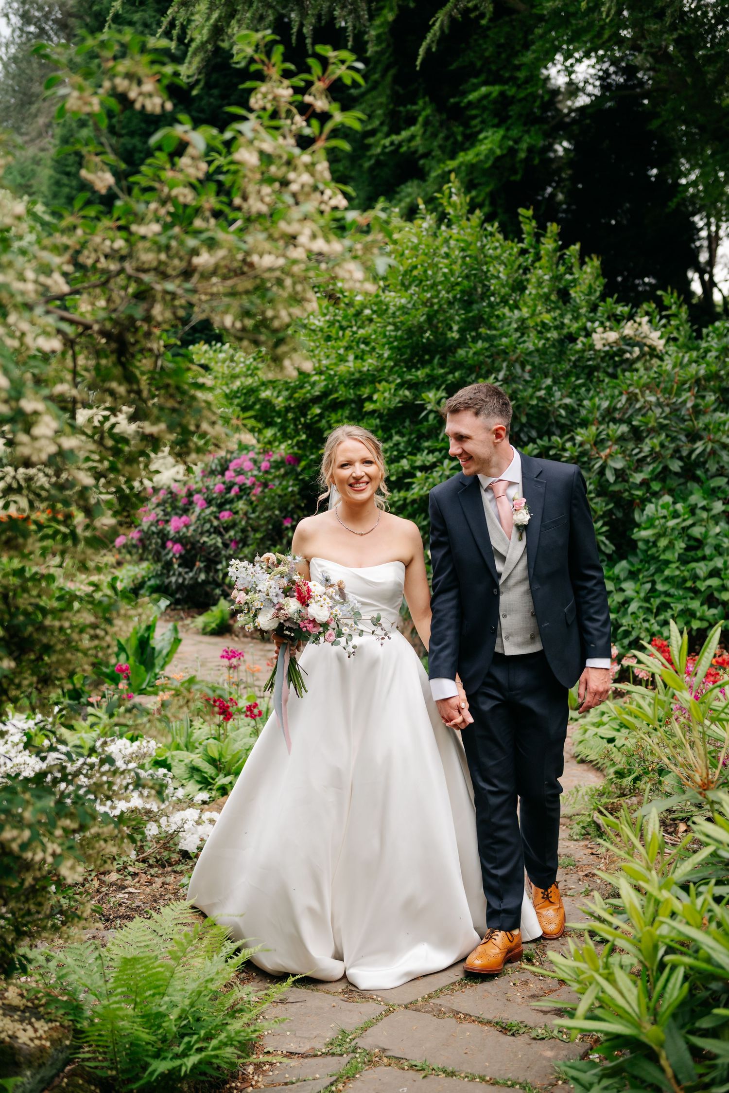 A bride and groom walk hand in hand through a lush garden, bride holding a floral bouquet in a strapless white gown.