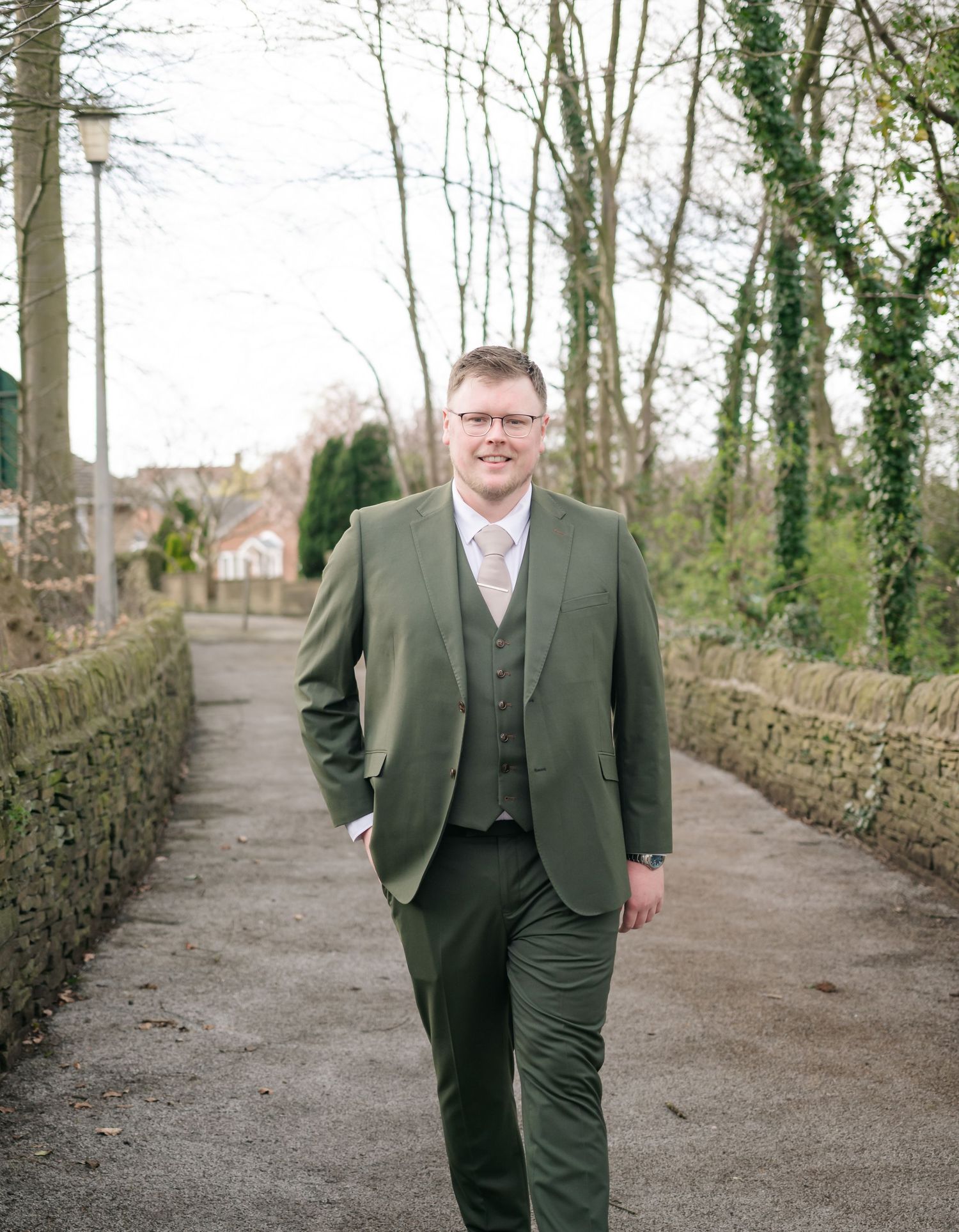 A person in a dark green three-piece suit walks along a tree-lined path with stone walls on either side.