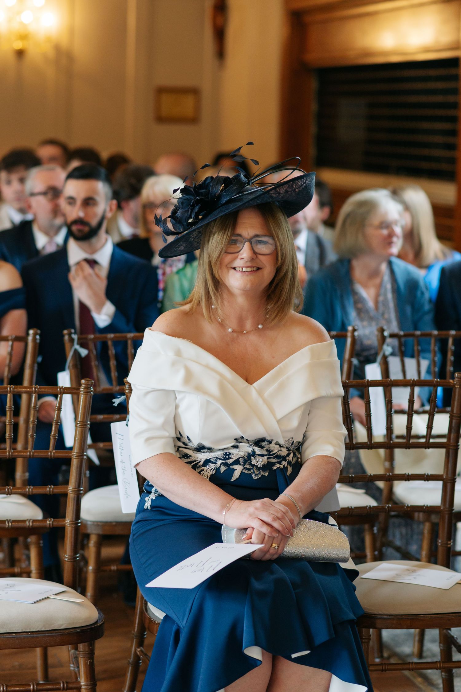 A wedding guest in a navy dress and elegant black fascinator sits in a ceremony venue.