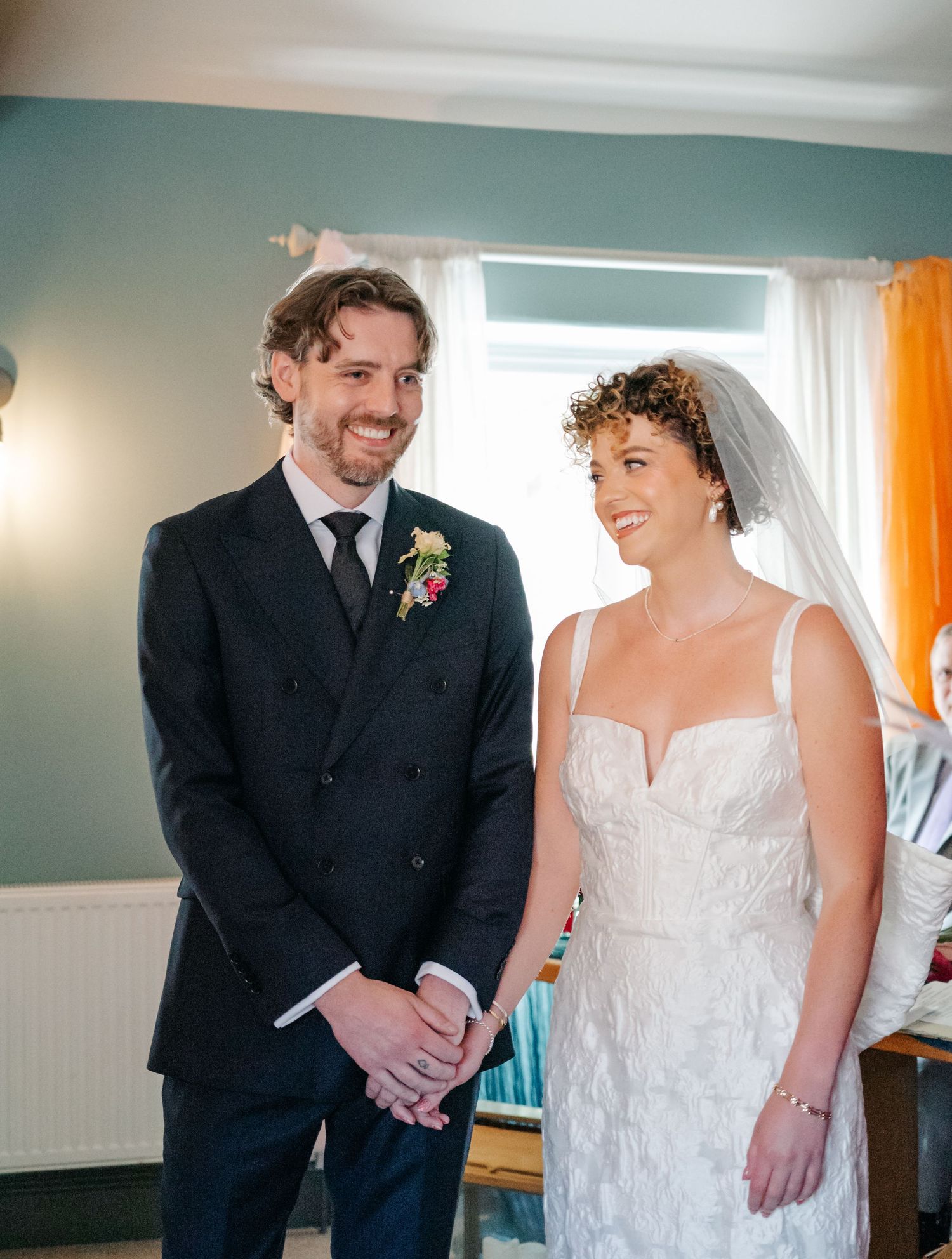 Couple shares intimate first look moment in bright indoor setting with orange curtains.