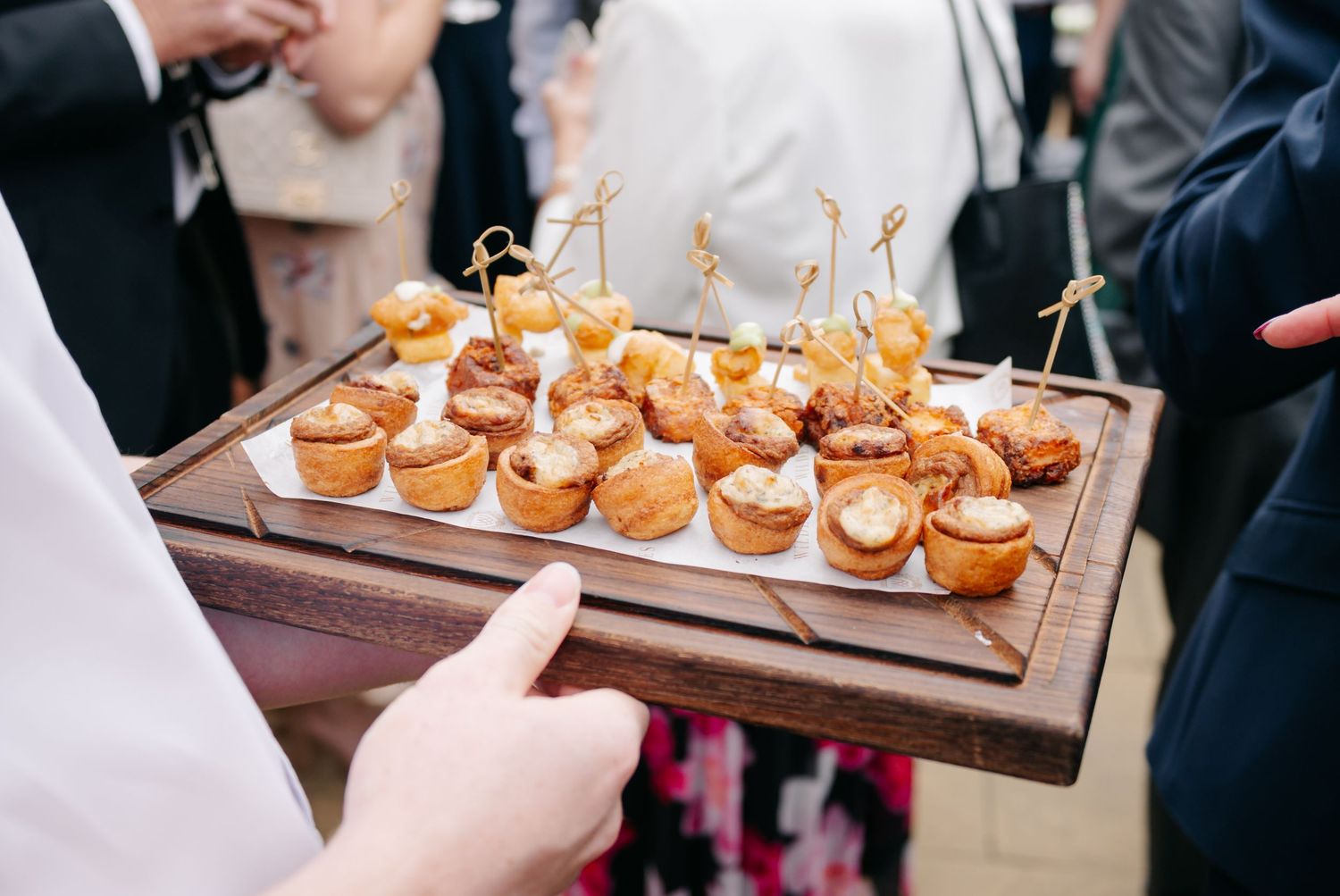 A wooden serving board displays mini sliders or appetizers with decorative toothpicks at an elegant catered event.