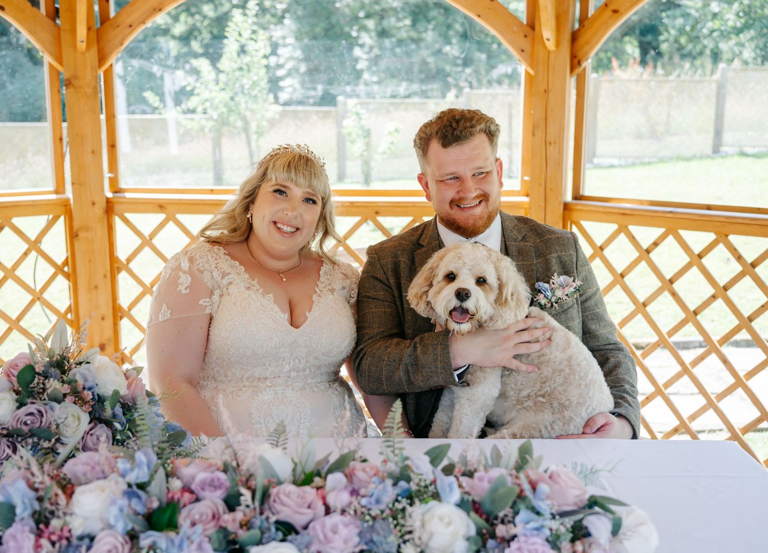 Bride and groom smiling with their fluffy dog at a wedding ceremony under a wooden lattice gazebo with pink floral arrangements.