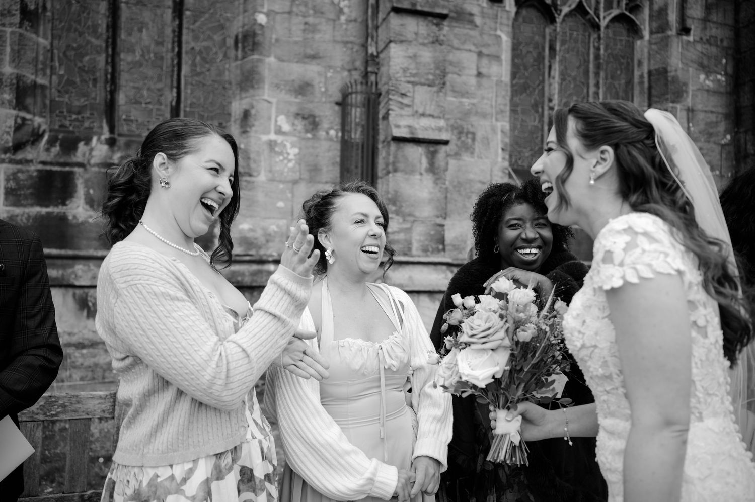Bride in a lace wedding dress holds a bouquet while laughing with three joyful women outside a historic stone building.