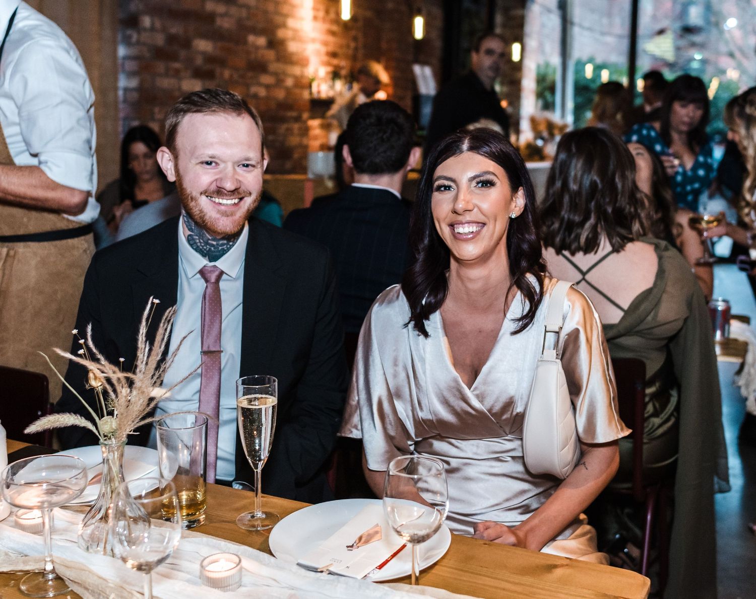 Wedding reception guests enjoying dinner at tables with brick wall backdrop.