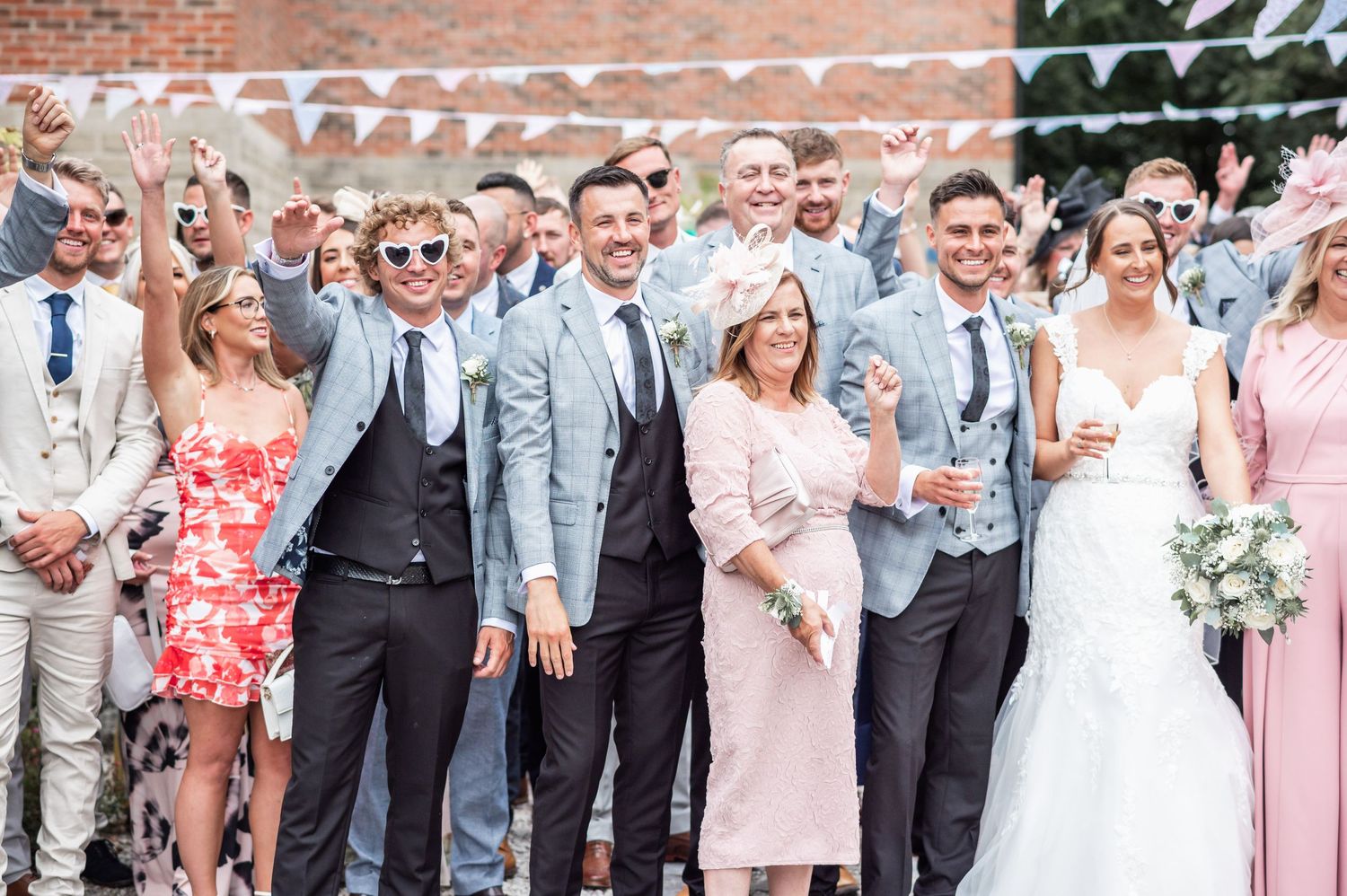 Wedding party and guests pose together outdoors against a brick wall background.