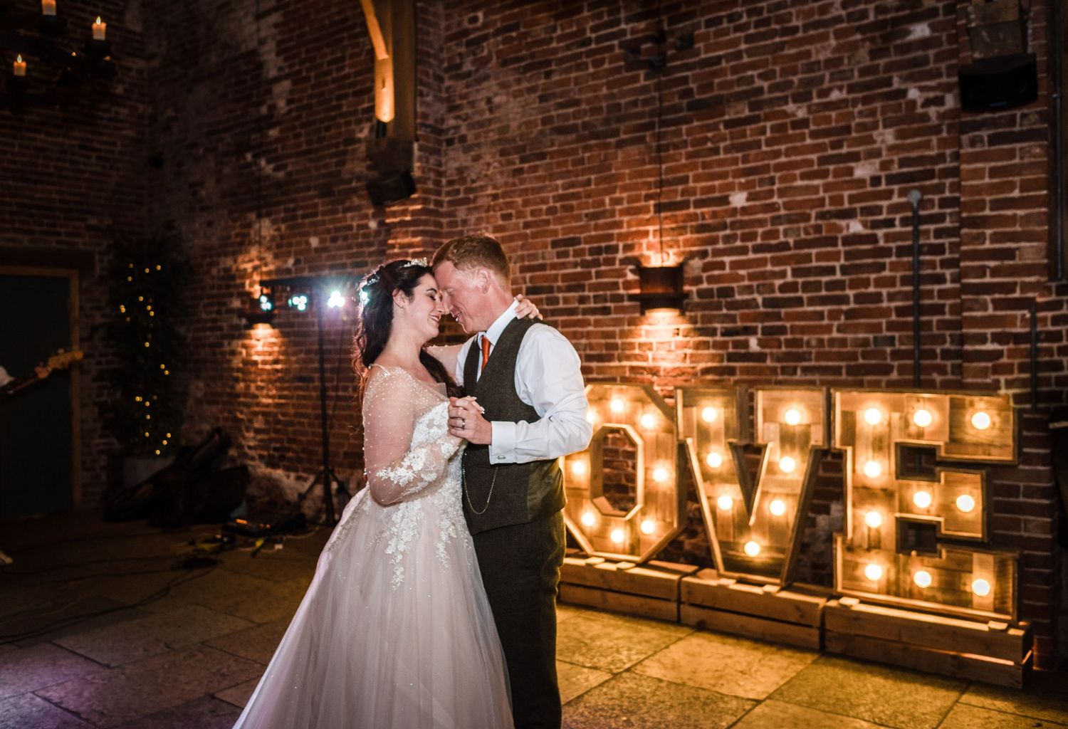A romantic dance moment near illuminated LOVE letters against exposed brick walls.