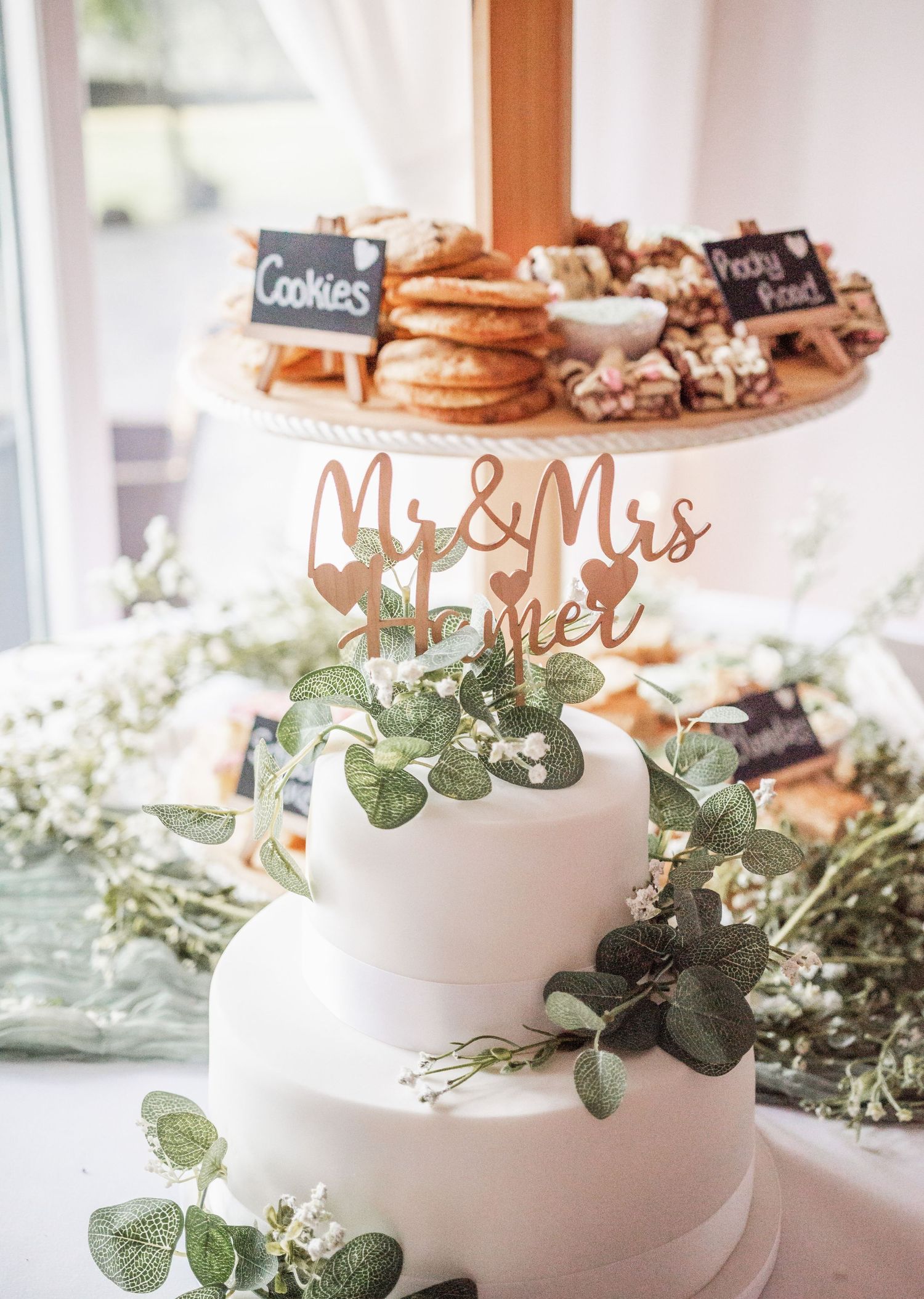 White two-tier wedding cake decorated with eucalyptus and a Mr & Mrs topper, surrounded by cookies.