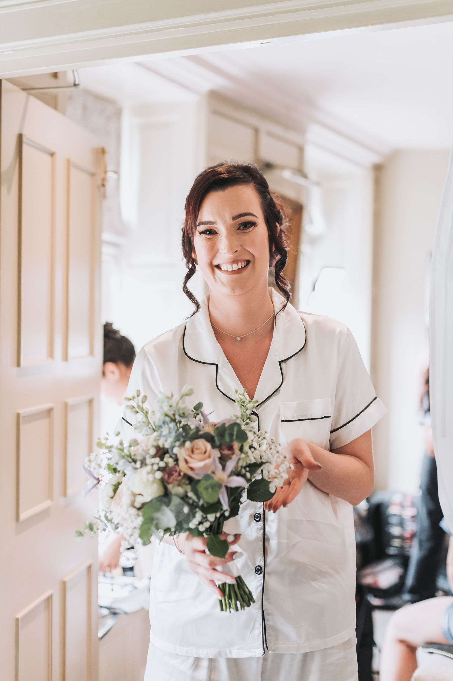 Bride in silk pajamas holds delicate bouquet of flowers during wedding morning preparations.