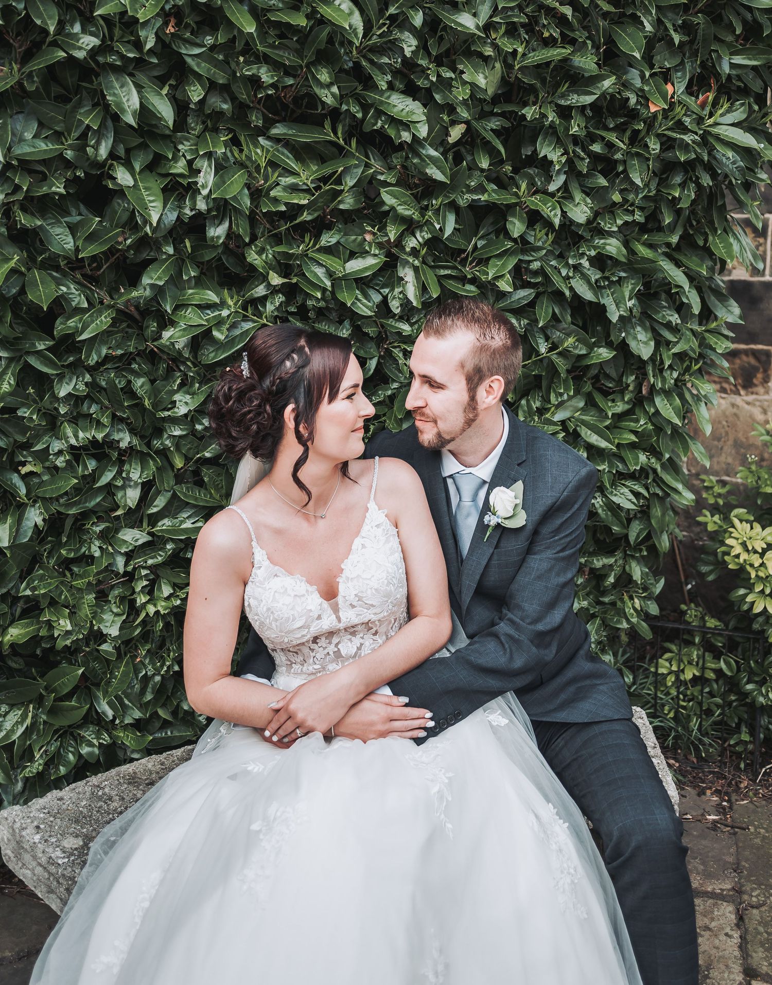 Romantic portrait of newlyweds sitting together against a lush green hedge background.