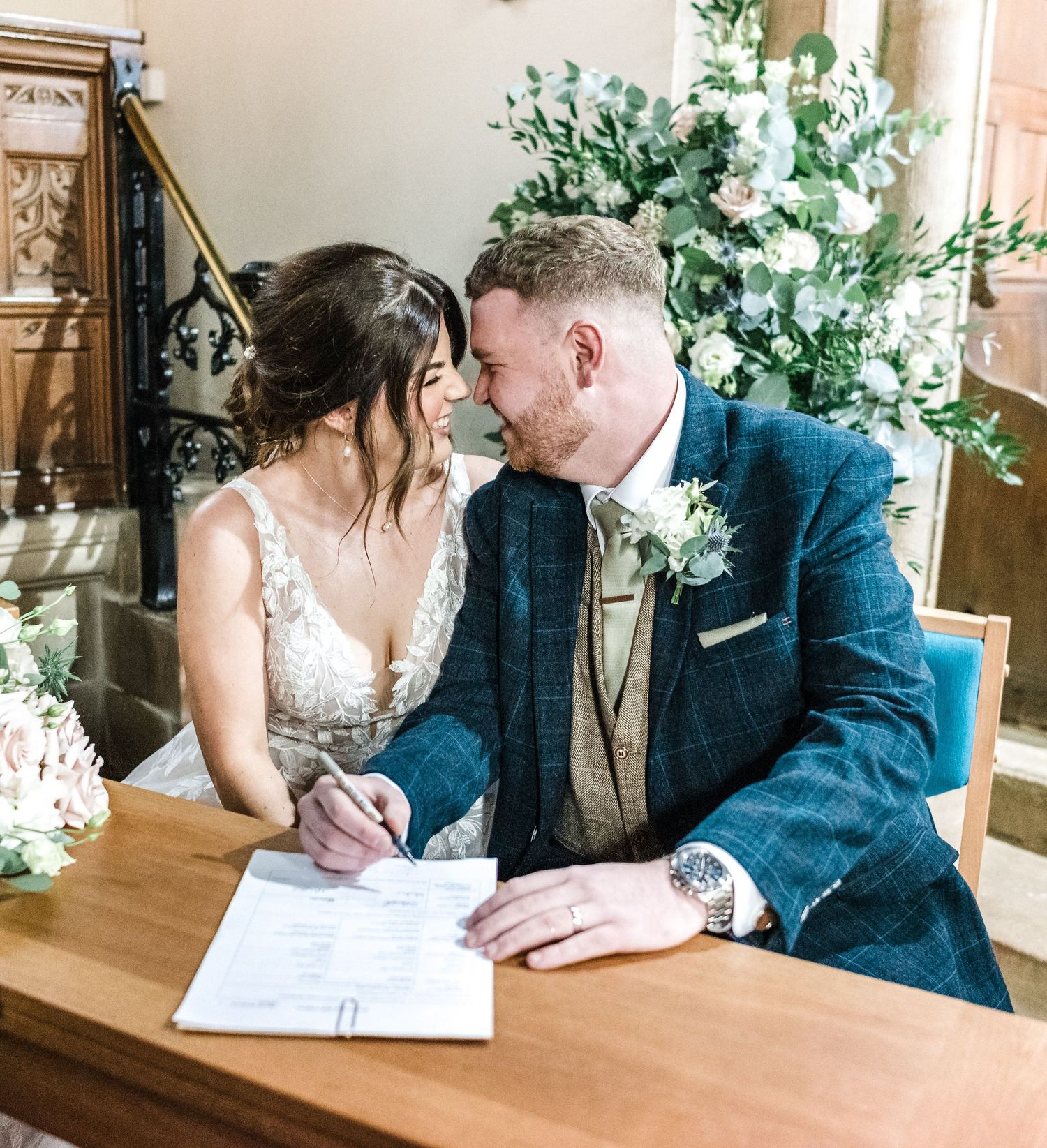 Wedding couple sharing tender moment while signing marriage documents at wooden desk.