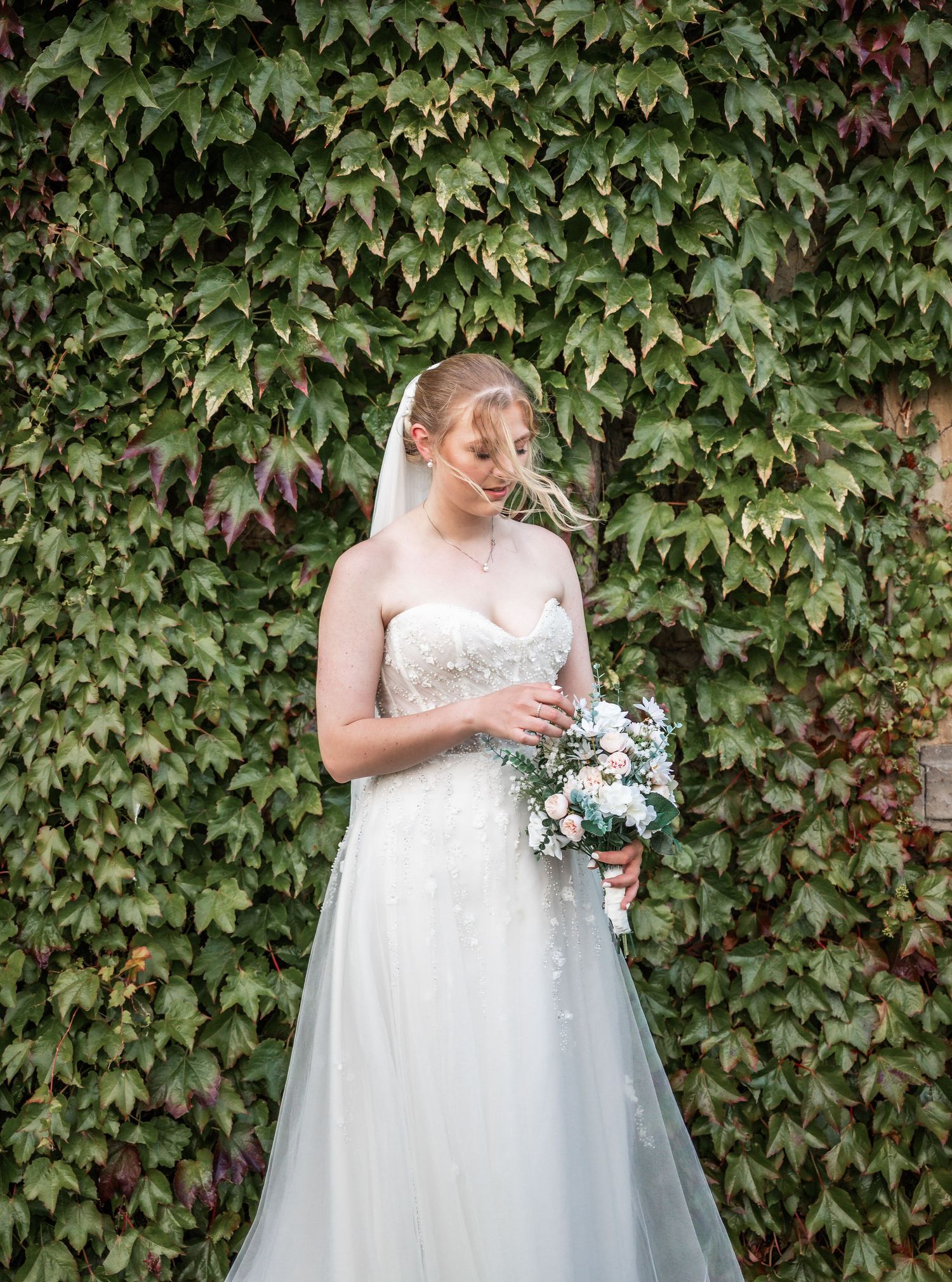 Elegant bride holding bouquet stands before ivy-covered wall in white lace wedding dress.