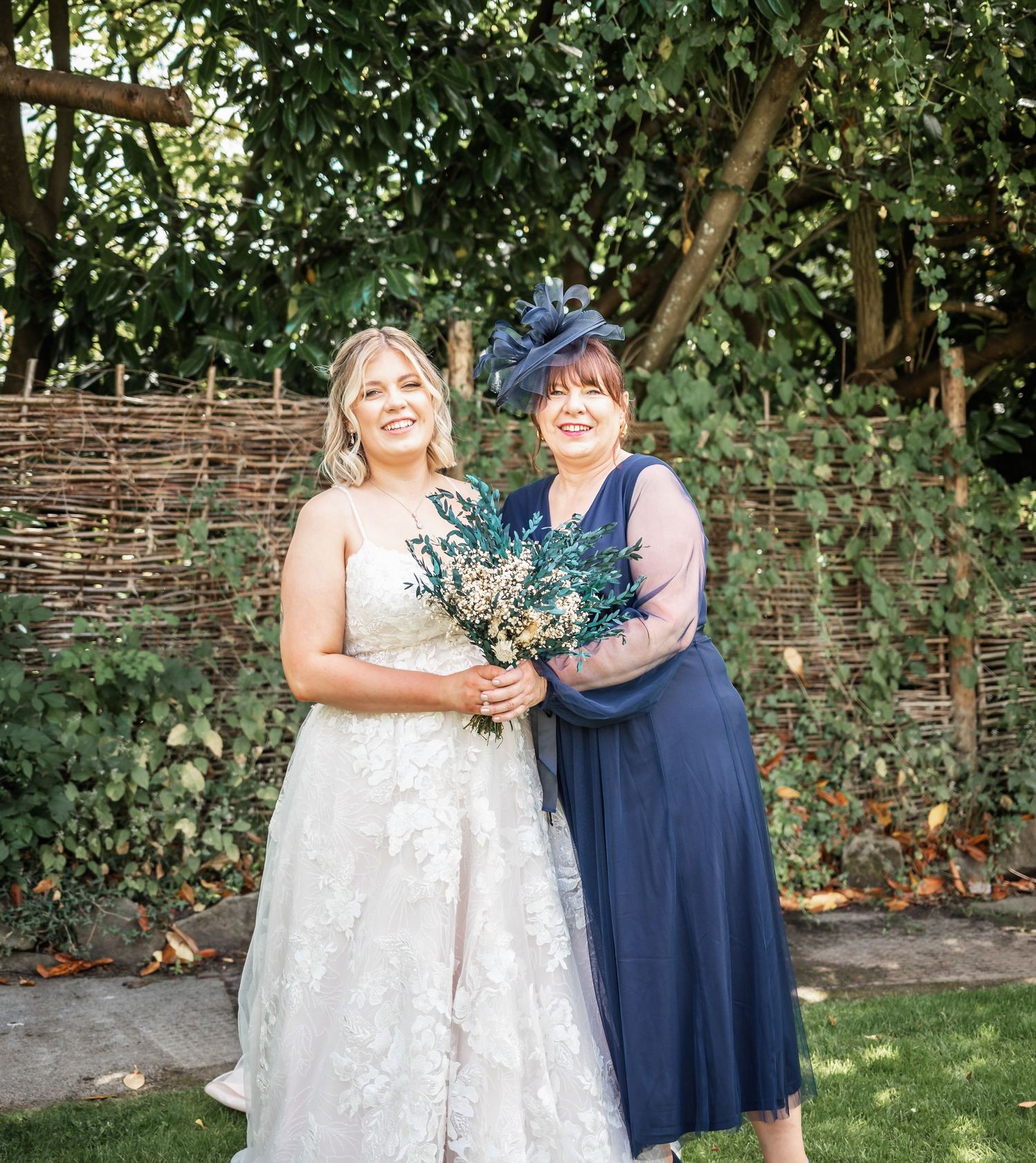 A white lace wedding dress and navy blue dress worn by pair in garden setting with rustic woven fence background.