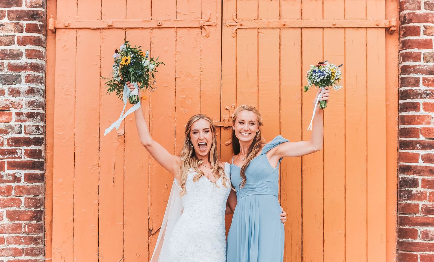 Bride and bridesmaid holding bouquets in celebratory pose against orange rustic door.