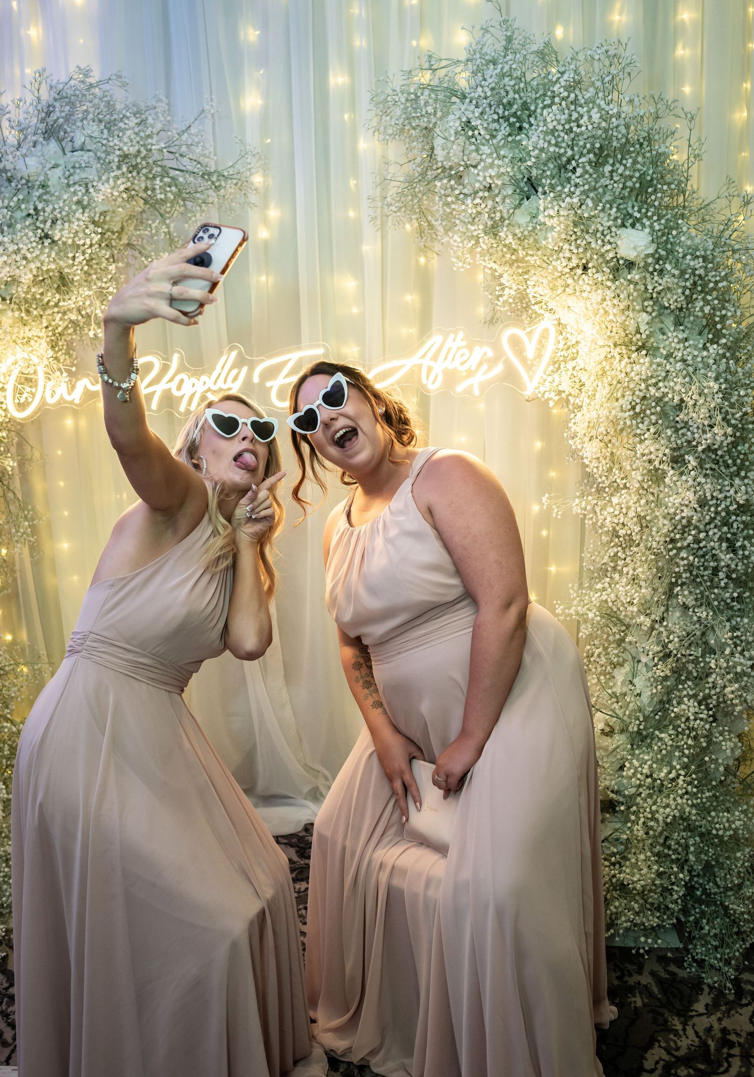 Two bridesmaids in champagne dresses taking selfie with funny expressions under neon sign.