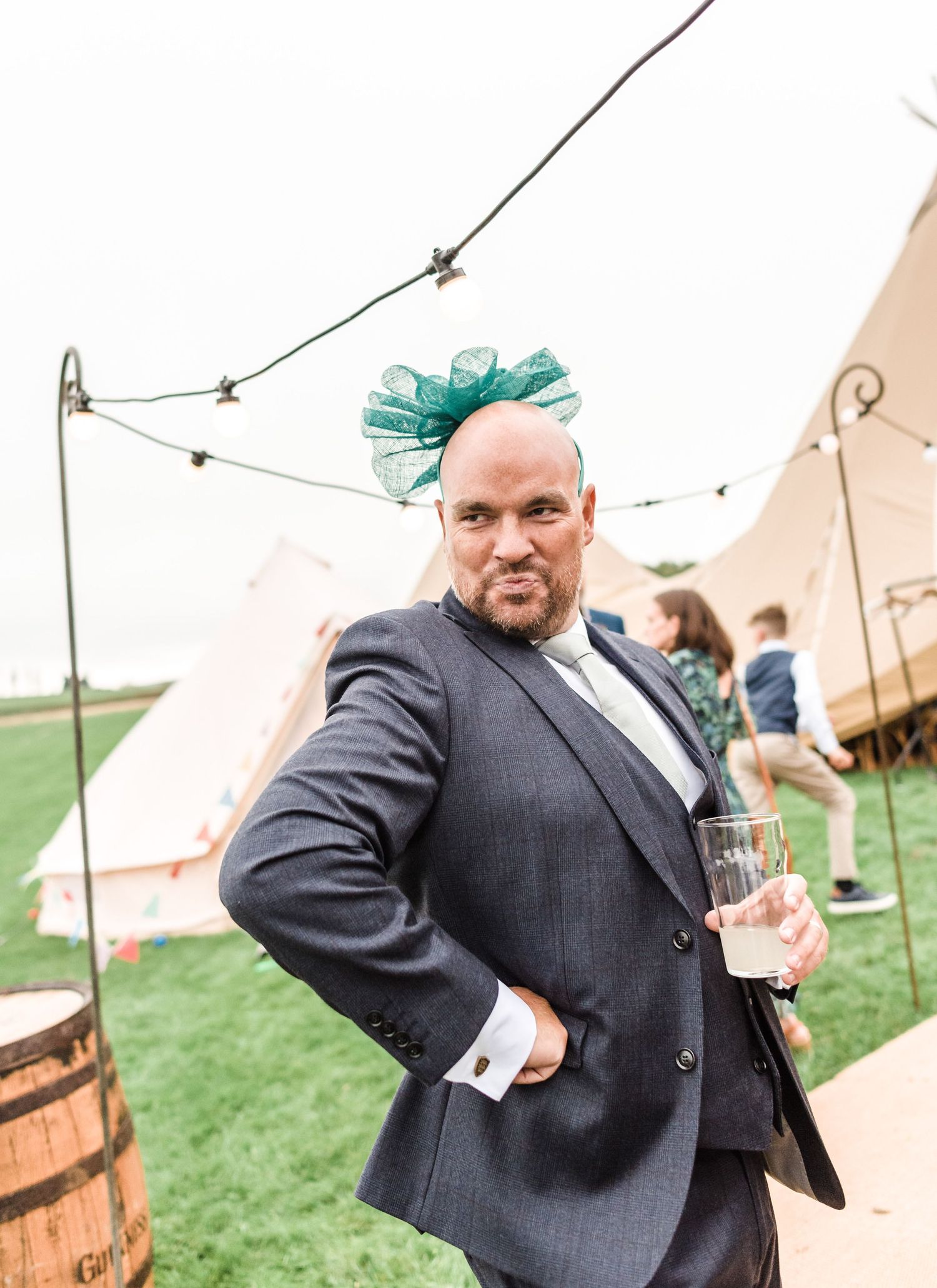 Man in gray suit wearing teal bow accessory poses with drink outside tent venue.