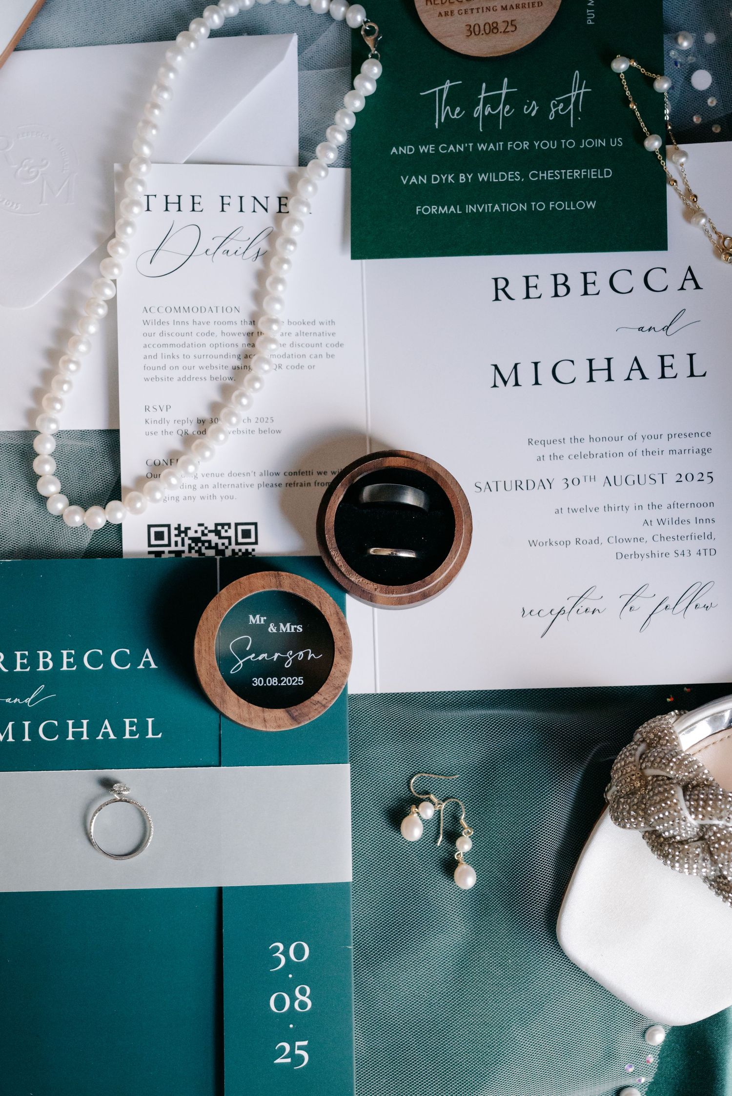 Wedding invitations, pearl necklace, and rings arranged on teal fabric with elegant typography.